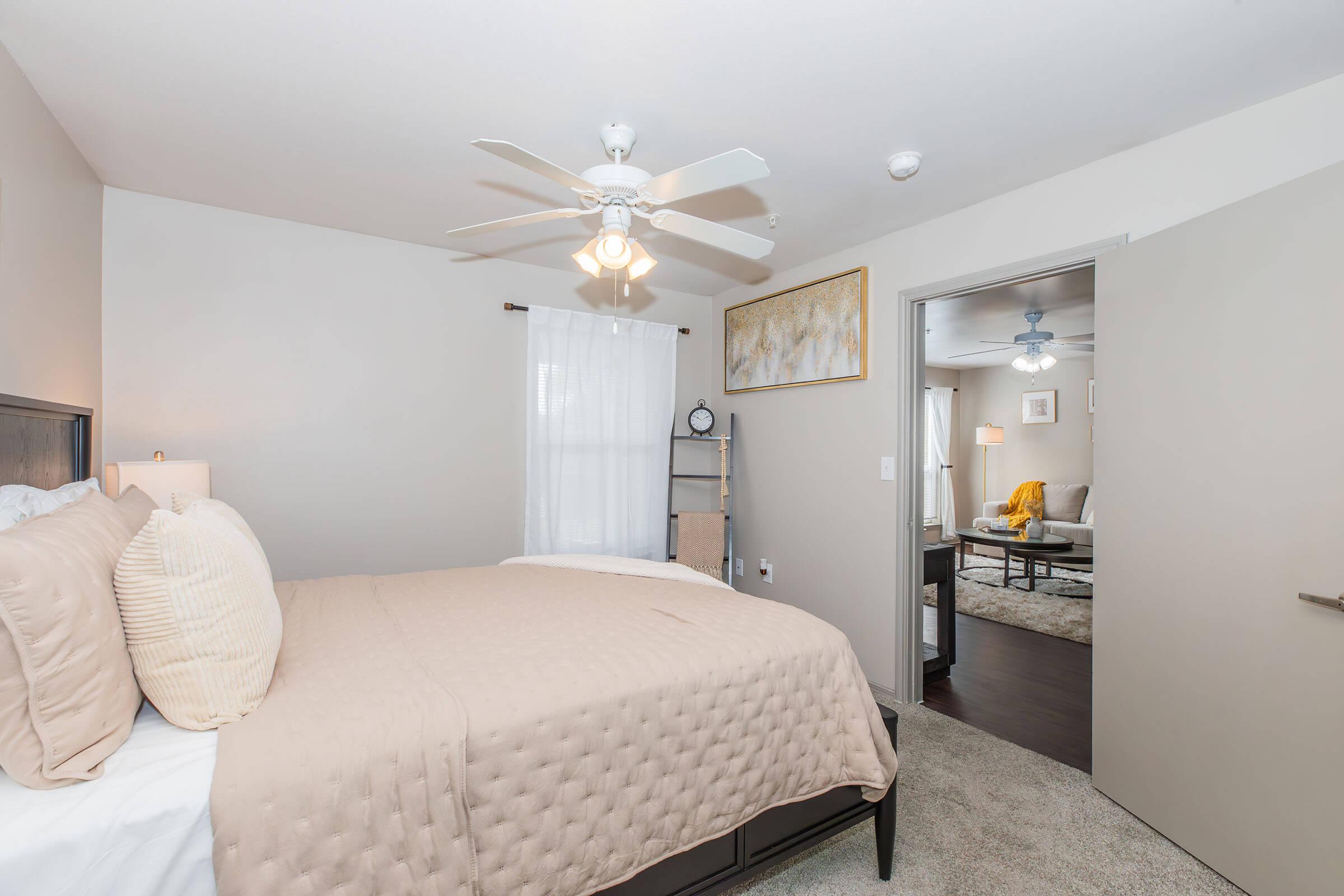 Cozy bedroom featuring a queen-sized bed with beige bedding, bedside lamps, and a ceiling fan. A window allows natural light and sheer curtains add privacy. A door leads to a living area with a yellow armchair, creating a warm and inviting atmosphere.