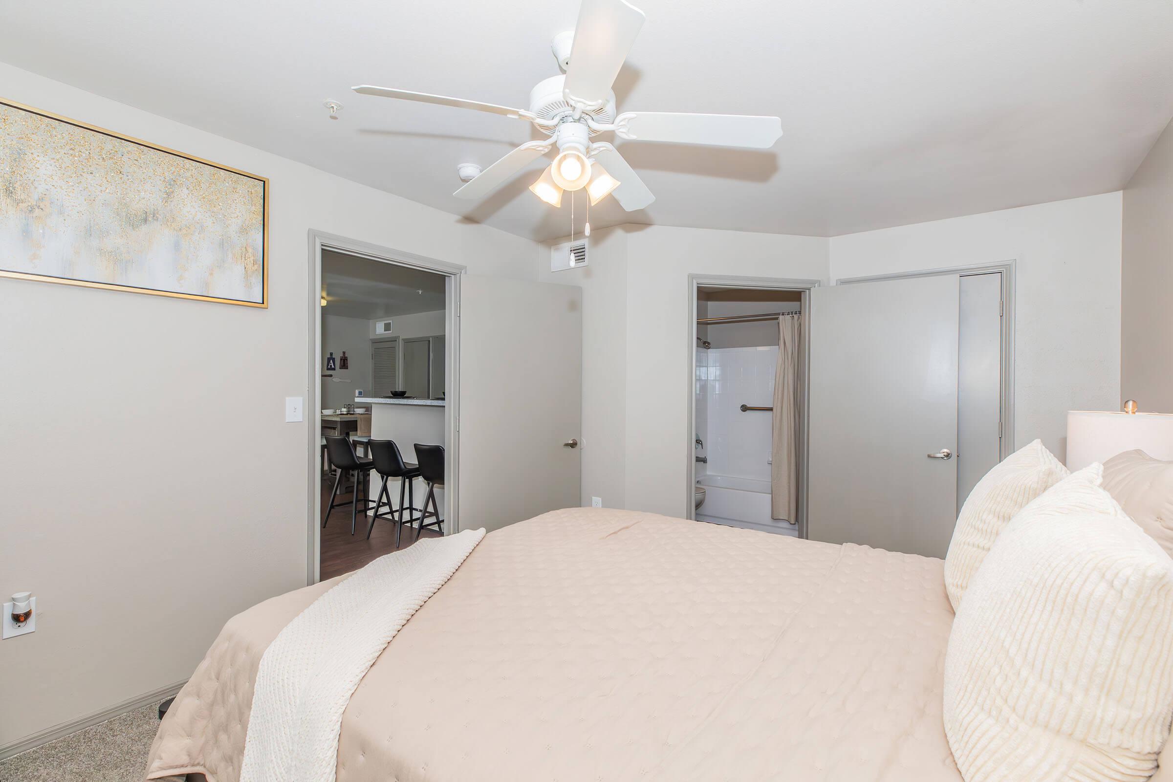 A cozy bedroom with a light beige bedspread and decorative pillows. There is a ceiling fan above, and a framed artwork hangs on the wall. Two doors lead to other areas, one likely to a bathroom with a visible shower curtain. The space is well-lit and inviting, with neutral-colored walls and carpet.