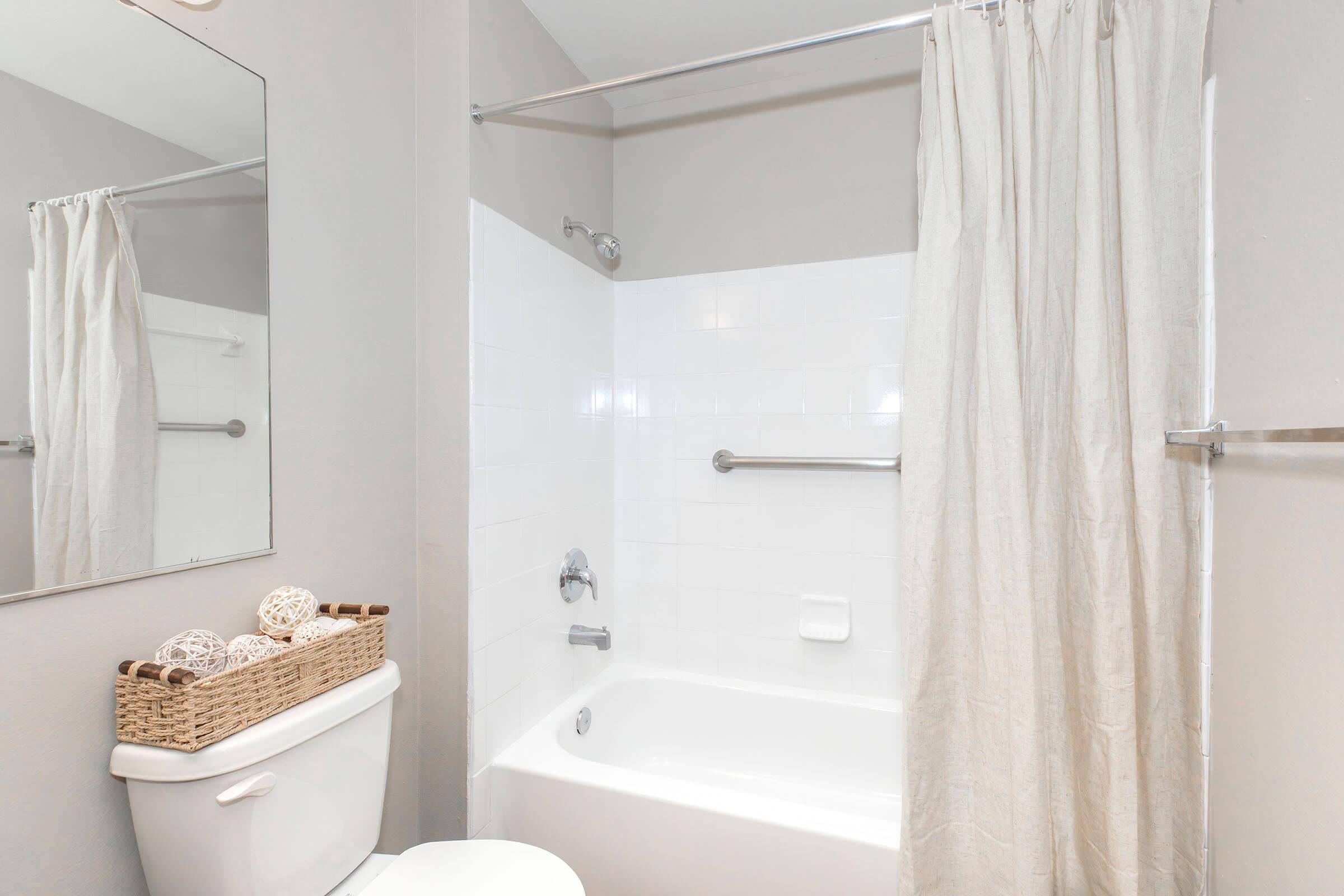 A clean bathroom featuring a white tub and shower with a light-colored curtain. A round mirror above a white toilet and a woven basket with decorative items are placed nearby. The walls are painted in a soft gray tone, creating a modern and minimalist ambiance.