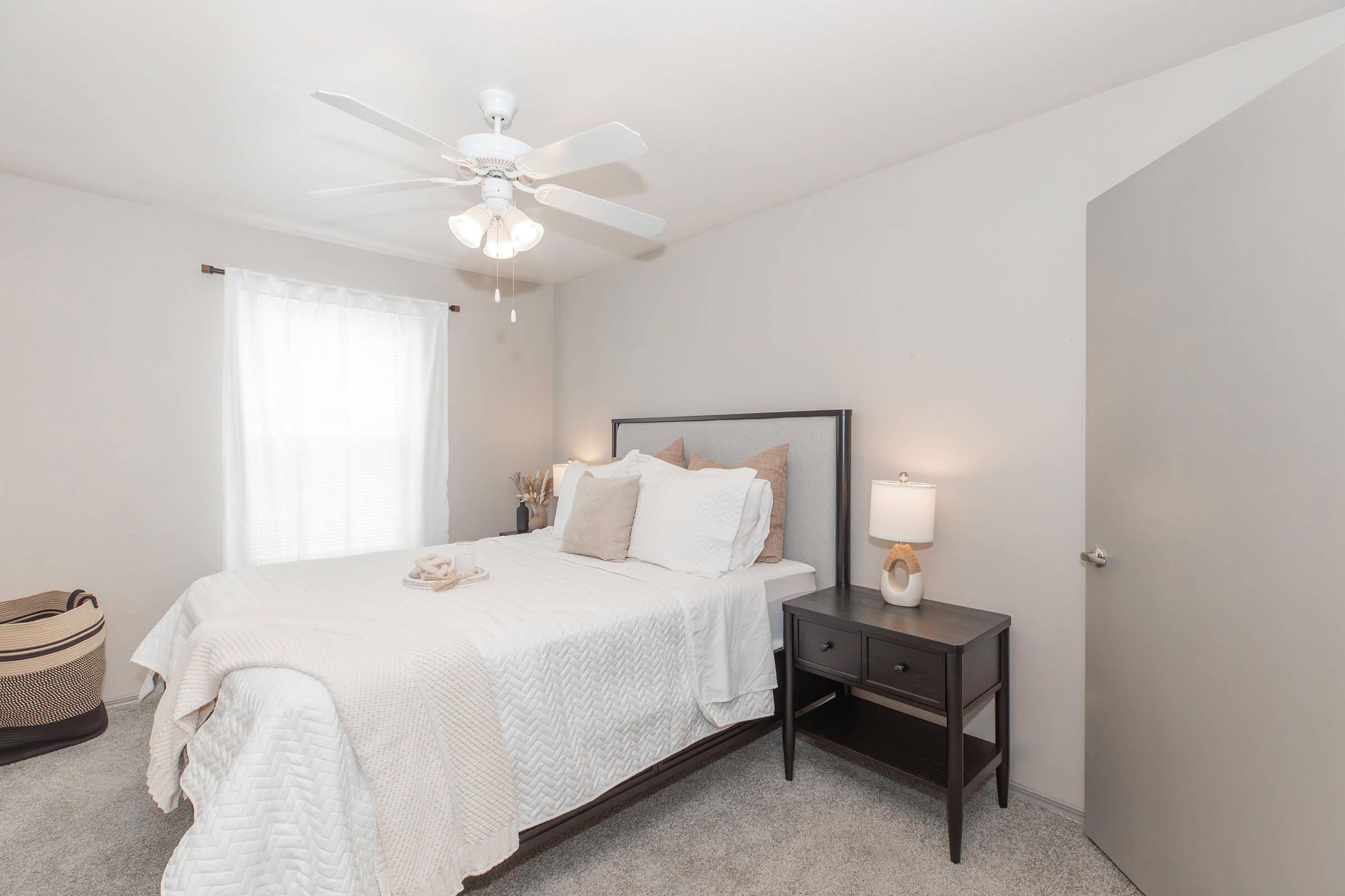 A cozy bedroom featuring a queen-sized bed with light bedding and decorative pillows. A wooden nightstand with a lamp sits beside the bed, and a ceiling fan is overhead. The room has a window with sheer curtains, allowing natural light, and a door leading to another space. A basket is placed on the floor.