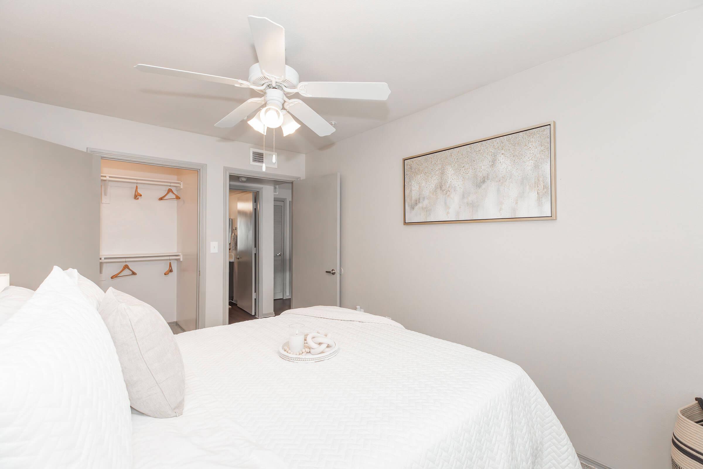 A bright bedroom featuring a queen-sized bed with white bedding and decorative items on top. A ceiling fan is visible, along with a closet in the background. The walls are painted a light gray, and abstract artwork hangs above the bed, adding a modern touch to the calm atmosphere.