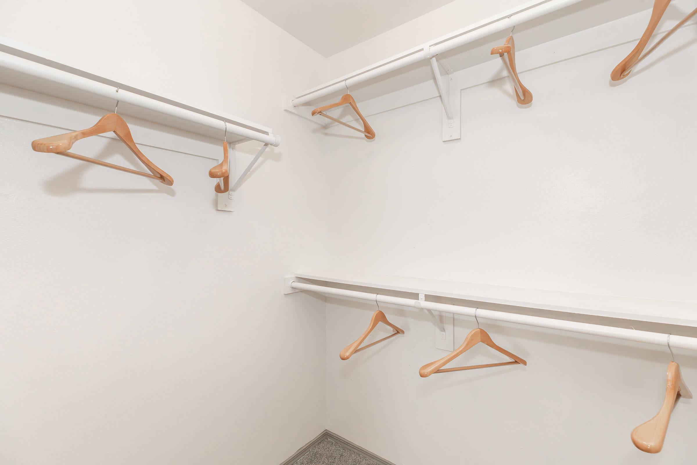 A minimalist closet with white walls, featuring several wooden hangers on white rods, and a shelf beneath the hangers. The floor is visible, adding to the uncluttered appearance.