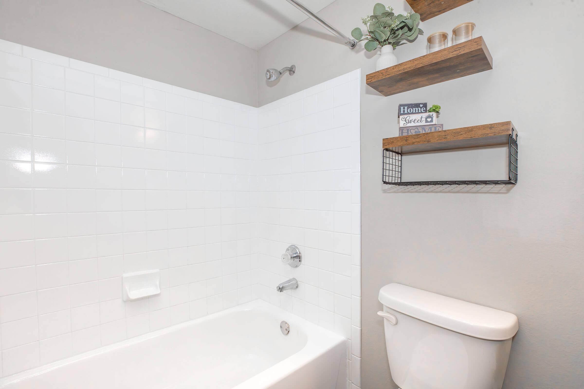 A clean, modern bathroom featuring a white tiled shower area with a bathtub, a toilet, and a wooden shelf above. The shelf holds decorative items and a small plant, adding a touch of greenery to the neutral-colored walls. Soft lighting enhances the bright and airy feel of the space.