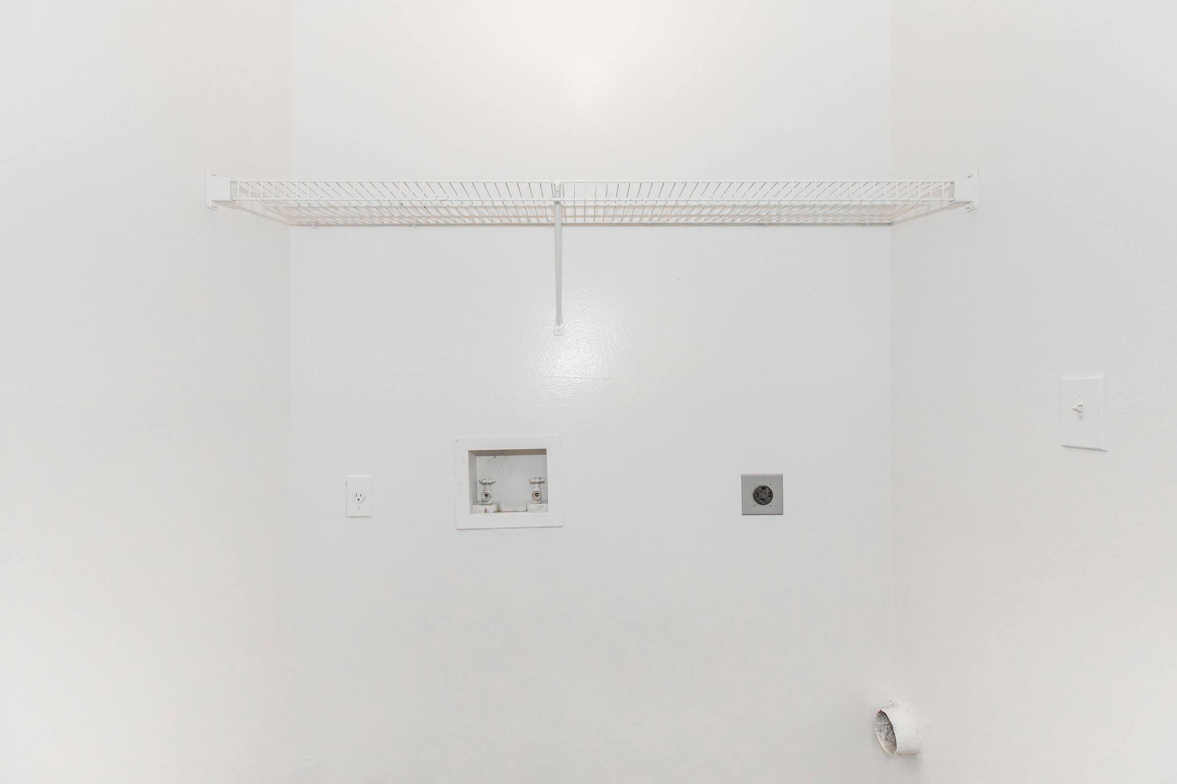 A blank white wall with a wire shelf mounted near the top. There are two electrical outlets and a small access panel visible on the wall. The floor is empty, and there is a vent near the bottom corner. The overall space appears clean and minimalistic, likely a laundry or utility area.