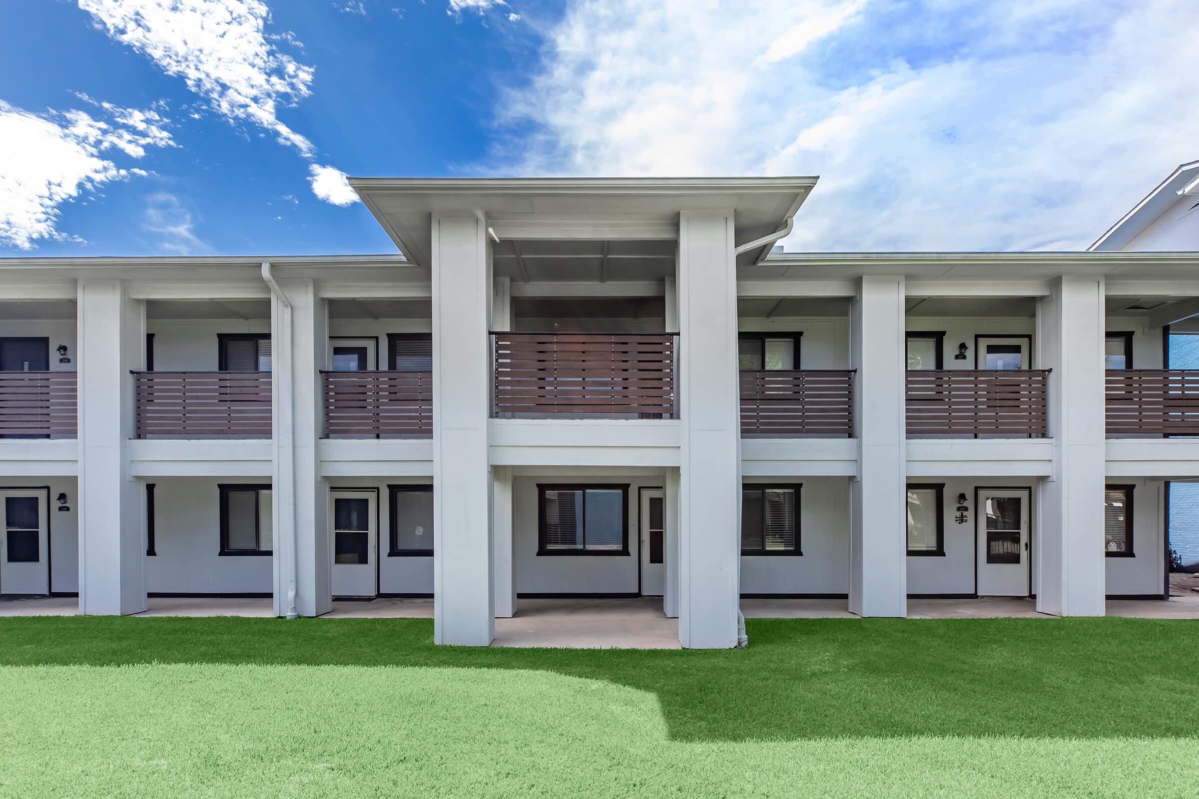 An exterior view of a modern two-story building featuring multiple apartment units. The facade includes large windows, balconies with wooden railings, and a grassy area in front. The sky is bright with clouds indicating a sunny day.