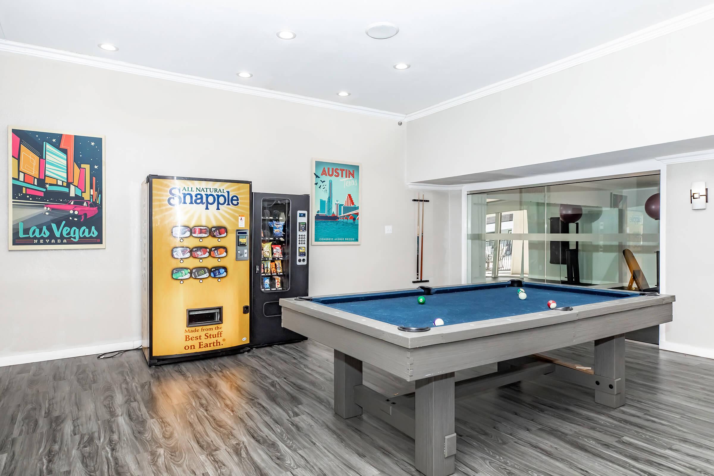 A recreational room featuring a blue pool table in the center, with a vending machine on the left displaying snacks and drinks. The walls are adorned with colorful posters of Las Vegas and Austin. The space is well-lit with recessed ceiling lights and has a modern, inviting atmosphere.