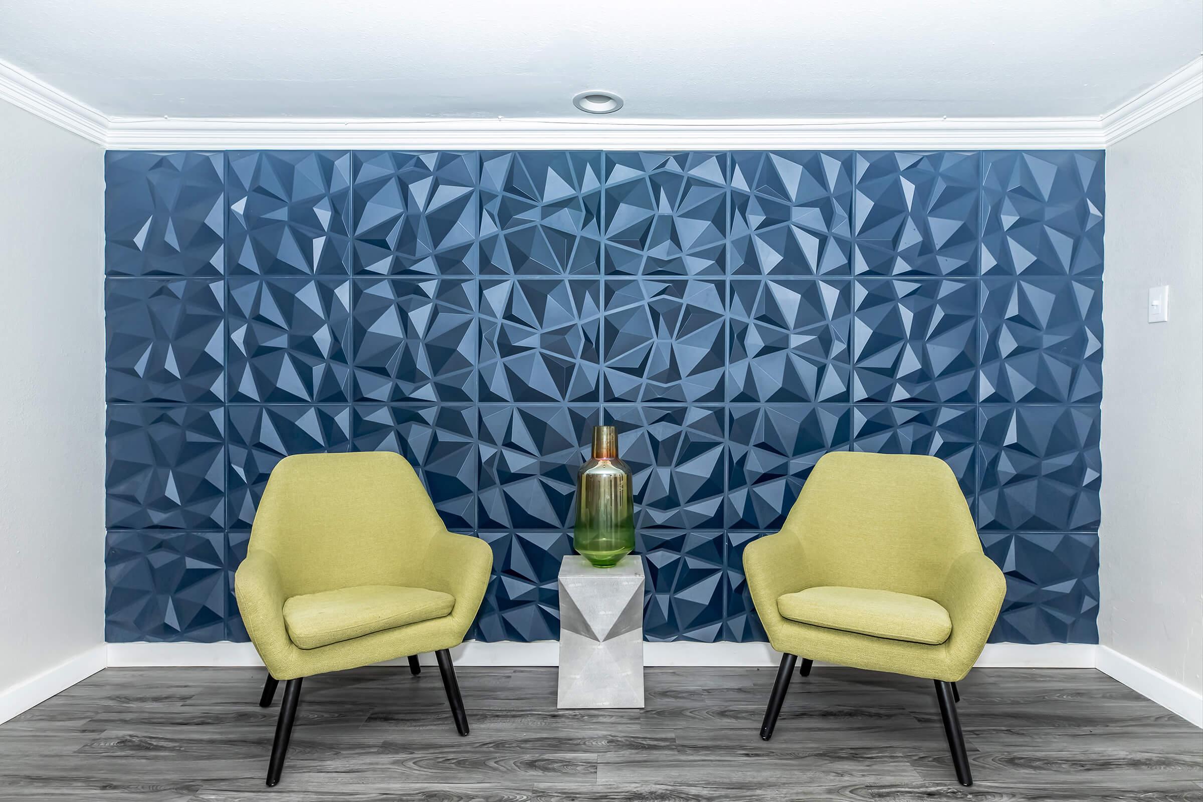 Two modern yellow chairs are positioned on either side of a geometric textured blue wall. Between the chairs is a sleek, silver side table holding a decorative glass bottle filled with green liquid. The flooring is a wood-like laminate, complementing the contemporary design.