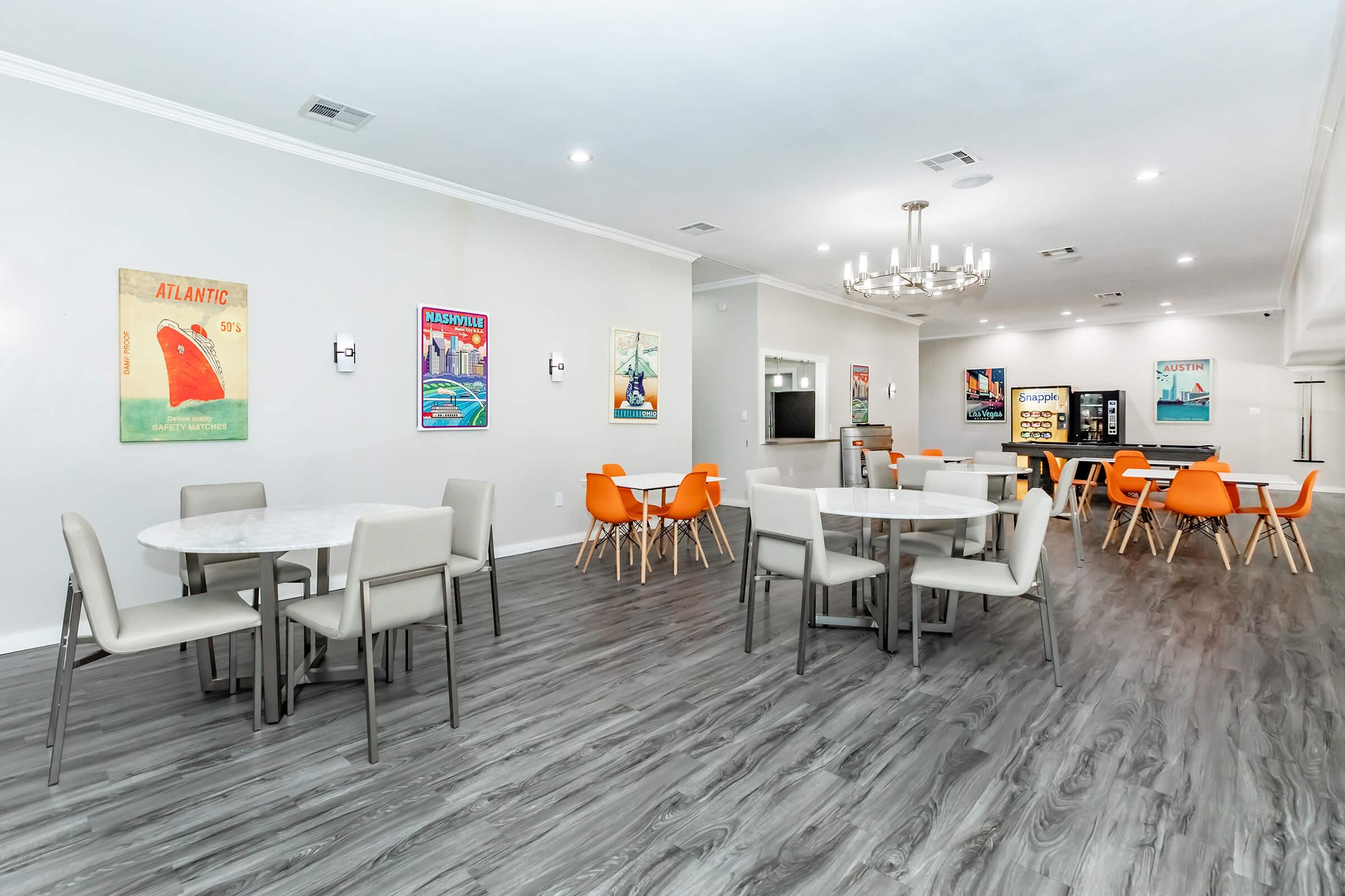 A modern dining area featuring several tables and chairs. The room has light-colored walls adorned with colorful vintage posters. There are two round tables surrounded by gray chairs and additional orange chairs at another table. The flooring is dark wood, enhancing the contemporary feel of the space.