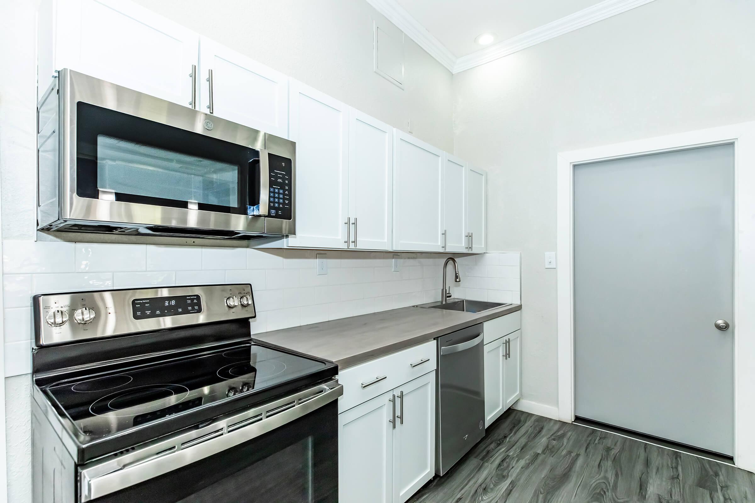 Modern kitchen featuring stainless steel appliances, including an oven, microwave, and dishwasher. The cabinetry is white with a sleek, minimalist design, and the countertop is a light gray. A gray door is visible on the right, complementing the contemporary aesthetic of the space.
