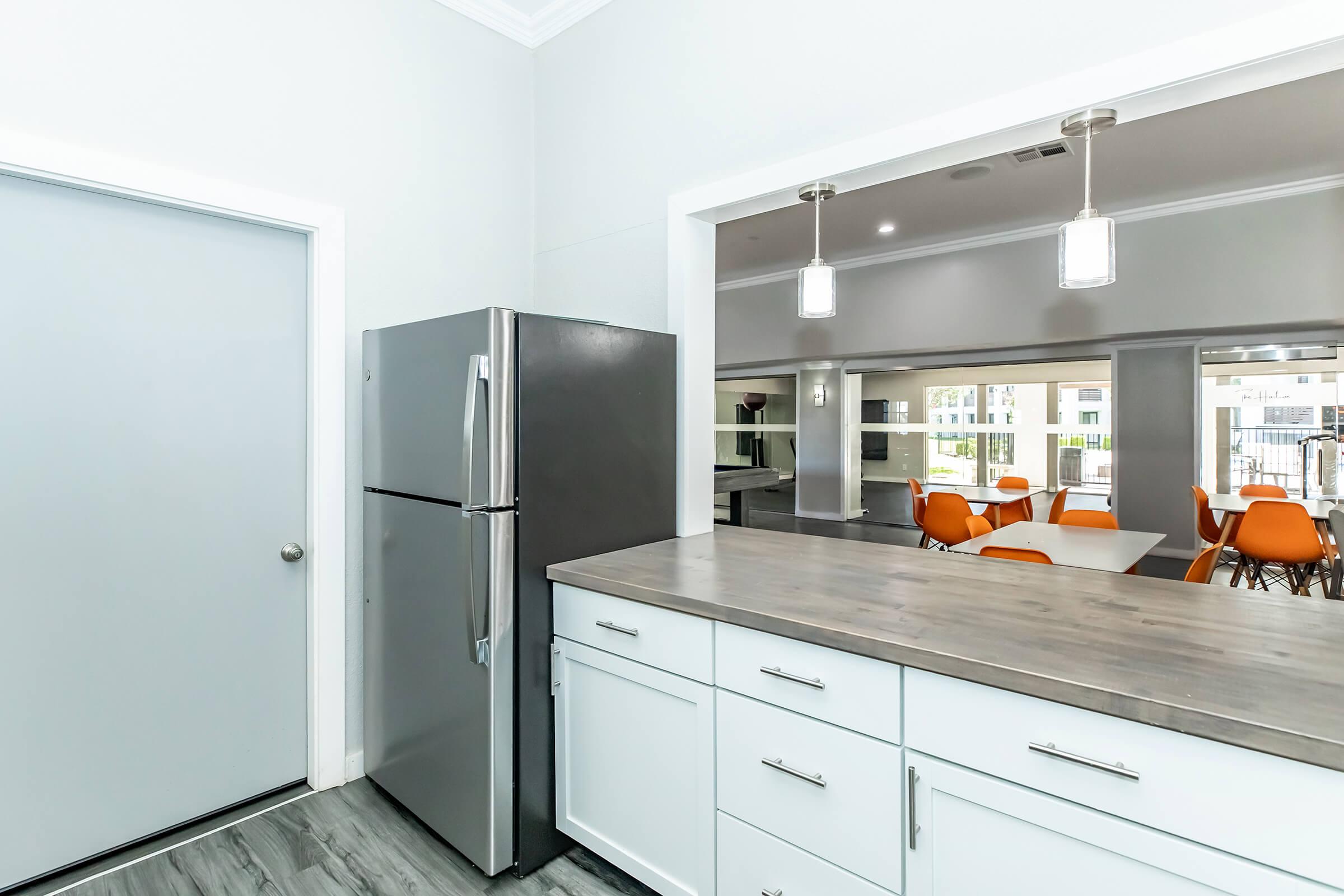 Modern kitchen setting featuring a silver refrigerator next to a light gray door. The countertop is a natural wood finish, and the background shows a dining area with orange chairs and tables. The space is well-lit, with pendant lights hanging above.