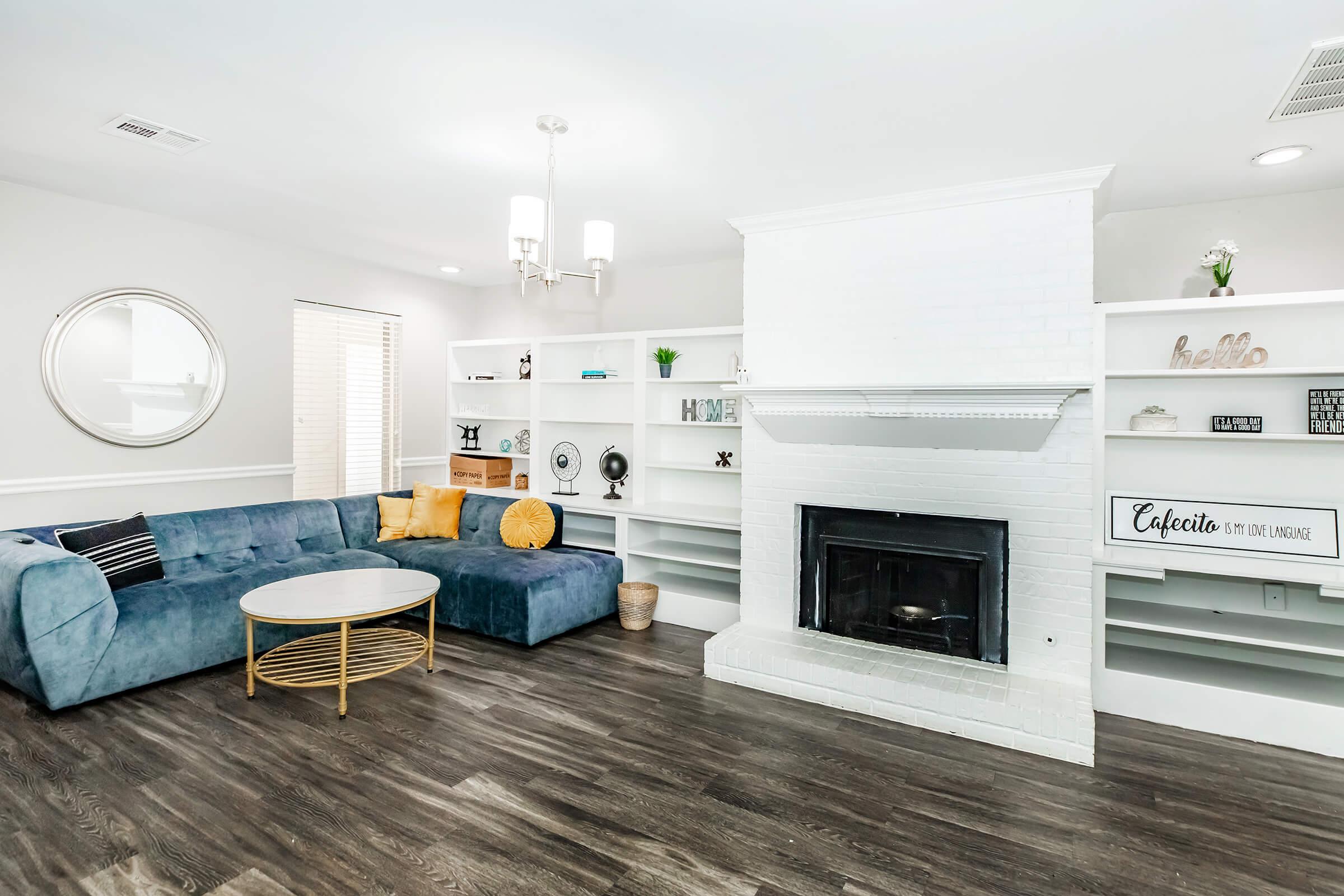 A modern living room featuring a blue sectional sofa with orange and yellow accent pillows, a round coffee table, and a white brick fireplace. Shelving units on either side of the fireplace display decorative items. A large mirror and a chandelier add to the bright, inviting atmosphere. The flooring is dark wood.