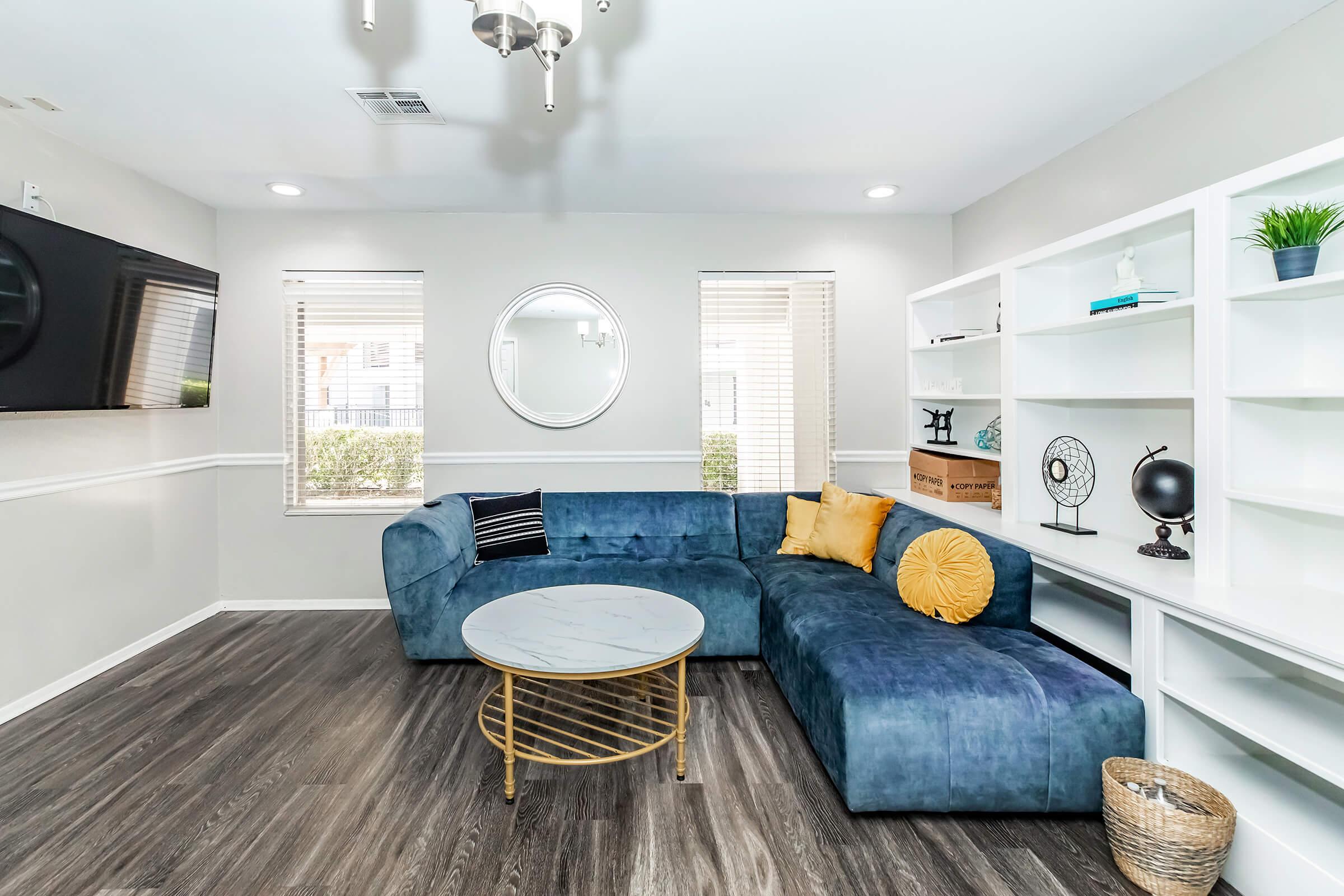 A modern living room featuring a blue sectional sofa with yellow throw pillows, a round marble-topped coffee table, and white built-in shelves. There is a large flat-screen TV mounted on the wall and natural light coming through windows, creating a cozy and inviting atmosphere.