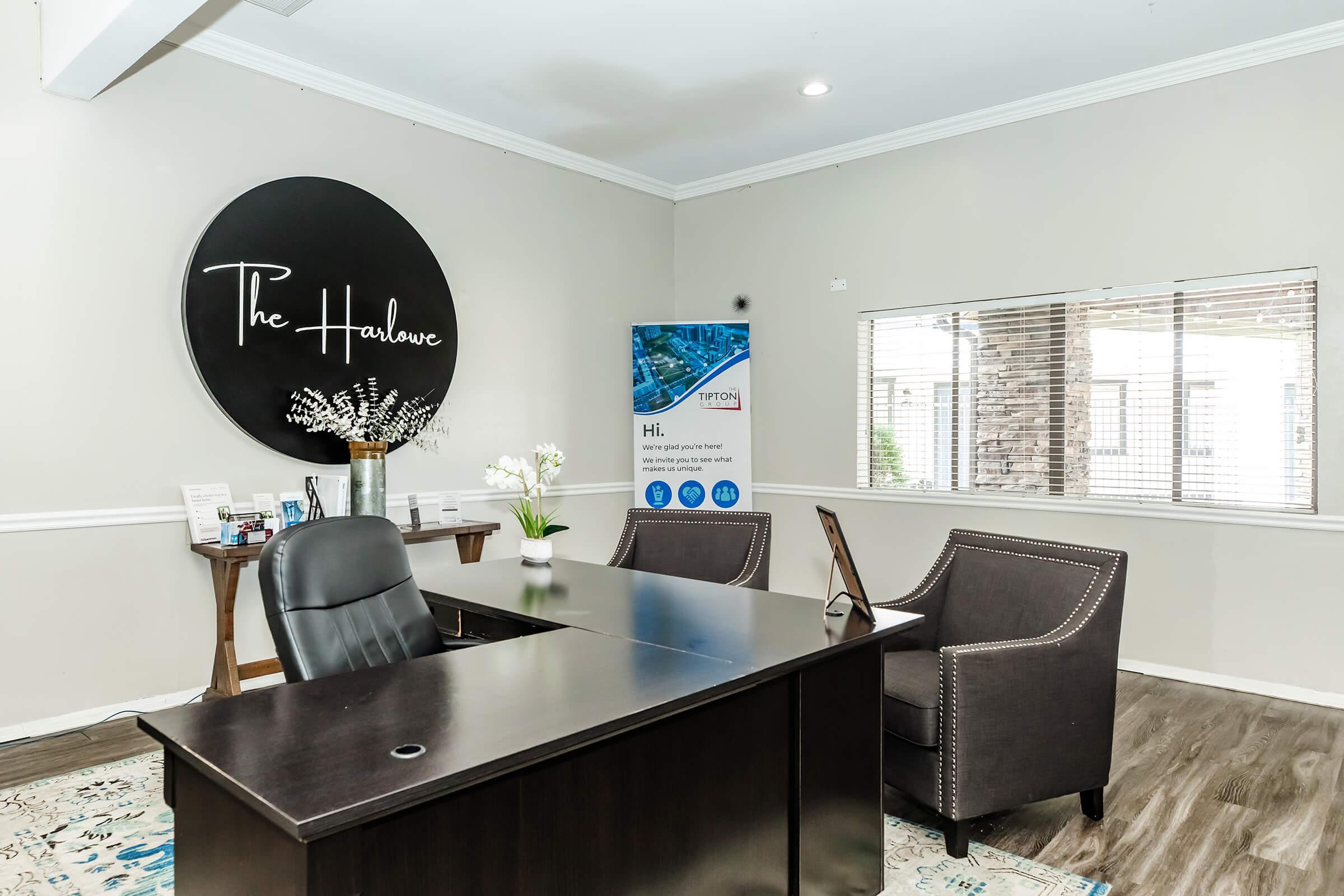 Reception area of an office featuring a round sign that reads "The Harlowe." A wooden desk is positioned in the center with a black office chair. There are two upholstered chairs alongside the desk. A window with blinds allows natural light, and there are decorative elements like a vase with flowers on the desk and promotional materials in the background.