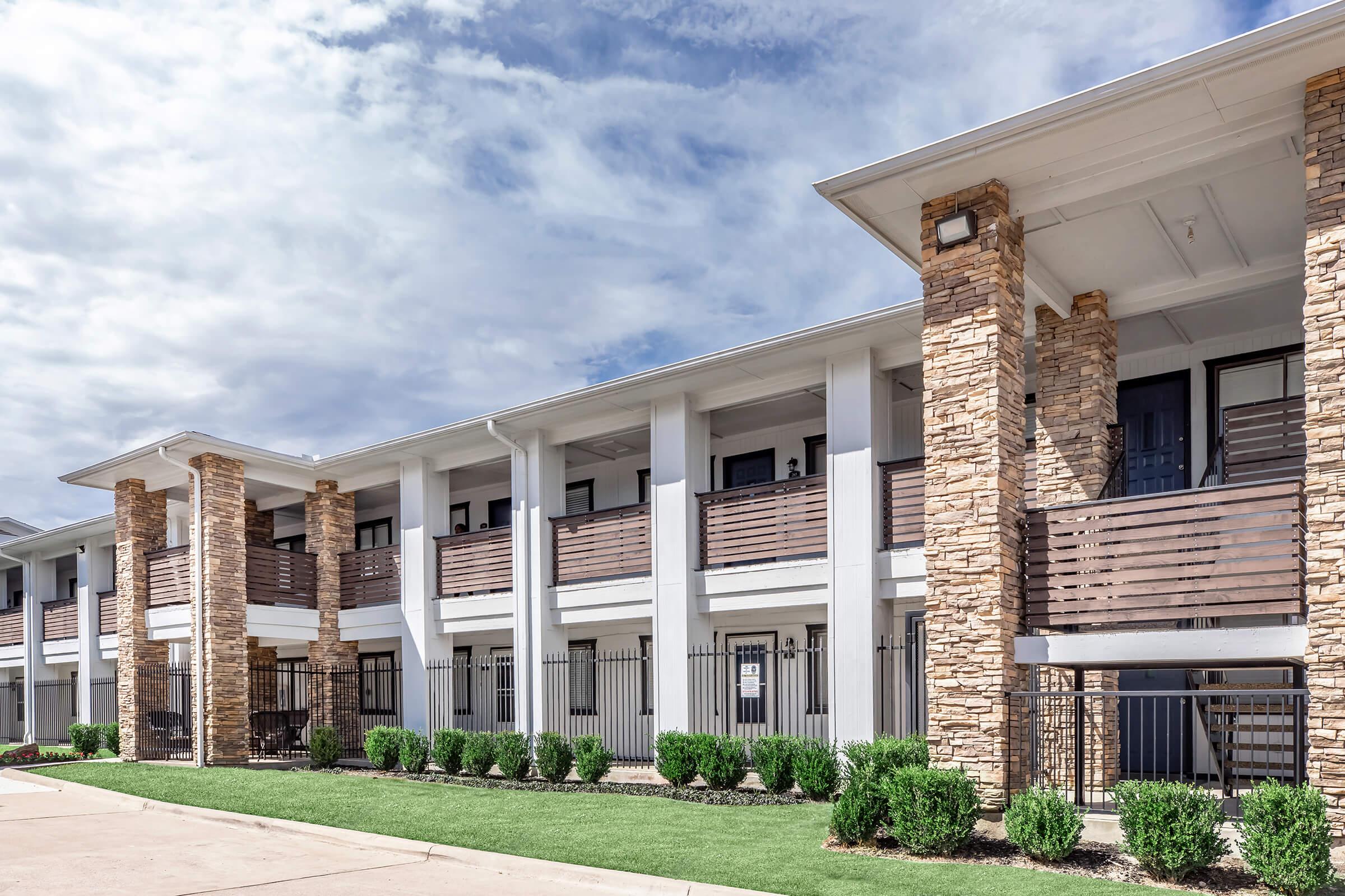A modern, multi-story building with stone and wood accents. The structure features multiple balconies with railings, surrounded by neatly trimmed shrubs. The sky above is partly cloudy, indicating a bright day. Nearby, a paved walkway leads to the entrances of the units.