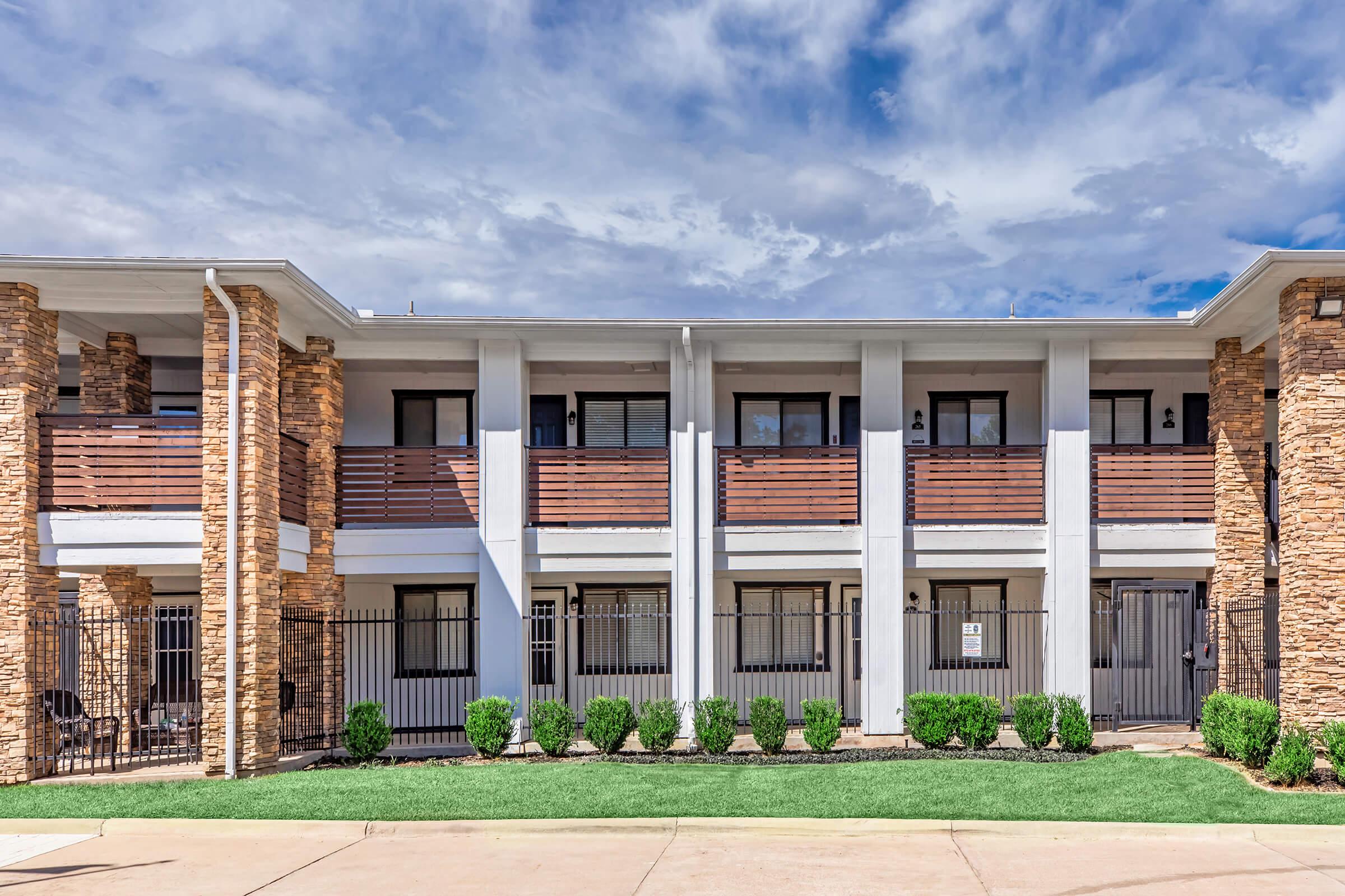 A modern two-story building with stone accents and wooden balconies. The exterior features neatly trimmed shrubs and a manicured lawn in front. The sky is bright with scattered clouds, creating a pleasant atmosphere. There are gated entries to the patios, enhancing security and privacy.