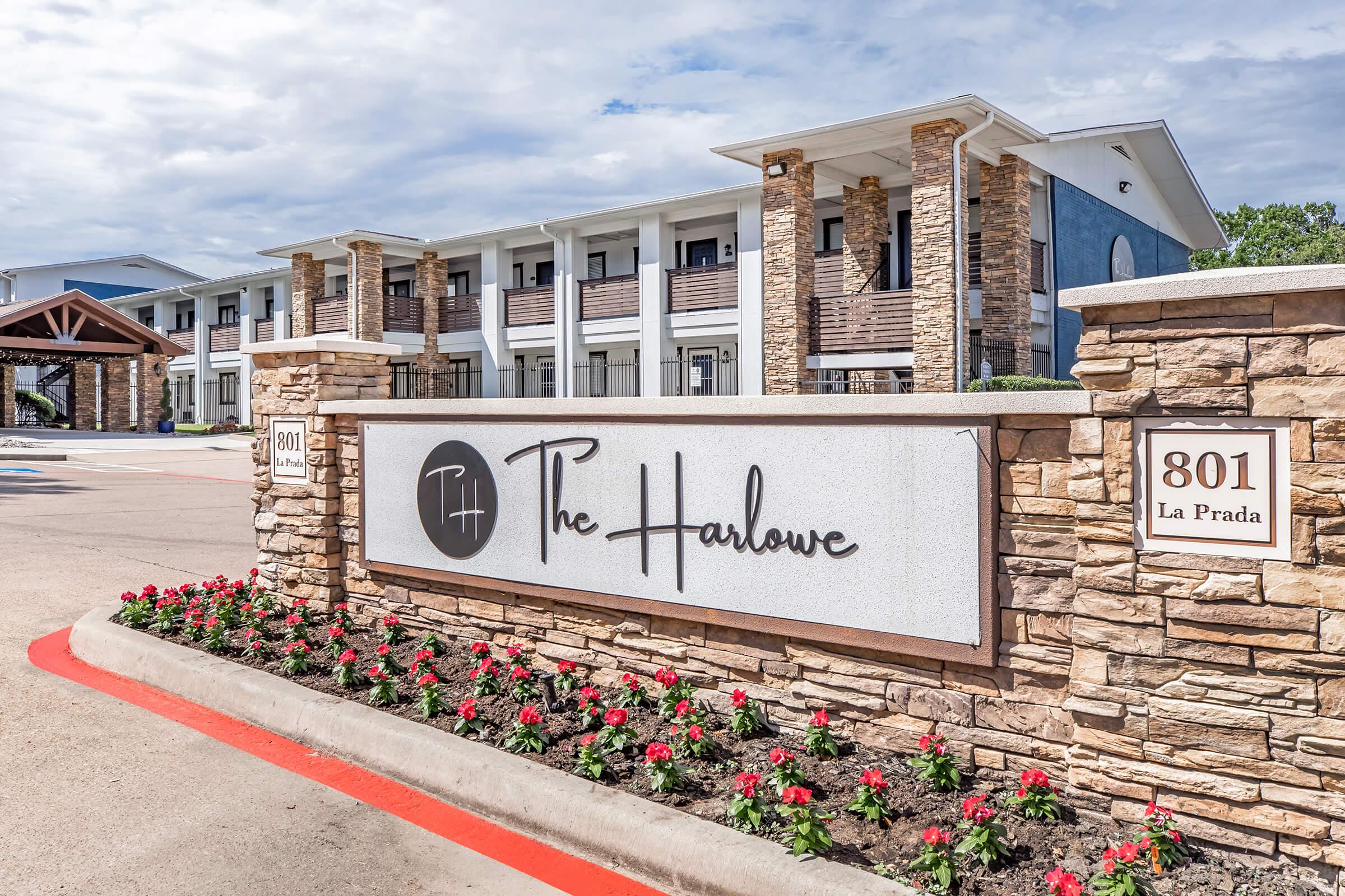 A well-maintained exterior view of The Harlowe, a multi-story building with a modern design. A prominent sign featuring the name "The Harlowe" is displayed in front, surrounded by vibrant landscaping with blooming flowers and a paved driveway. The sky is partly cloudy, contributing to a welcoming atmosphere.