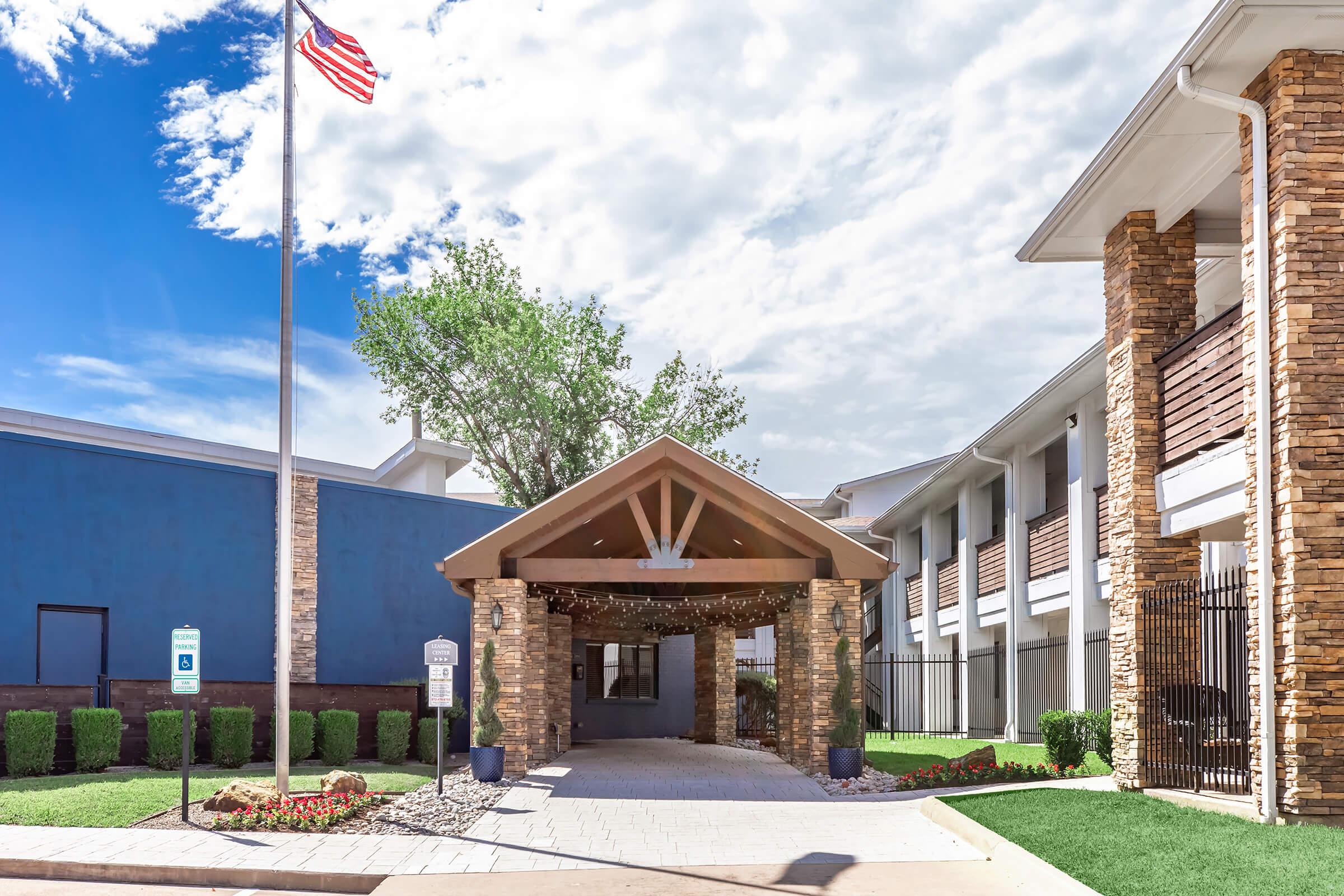 Entrance of a building featuring a covered awning supported by stone pillars, with decorative lights. In the background, a blue building is visible along with neatly trimmed hedges and flowers. An American flag flies on a pole to the left under a partly cloudy sky.