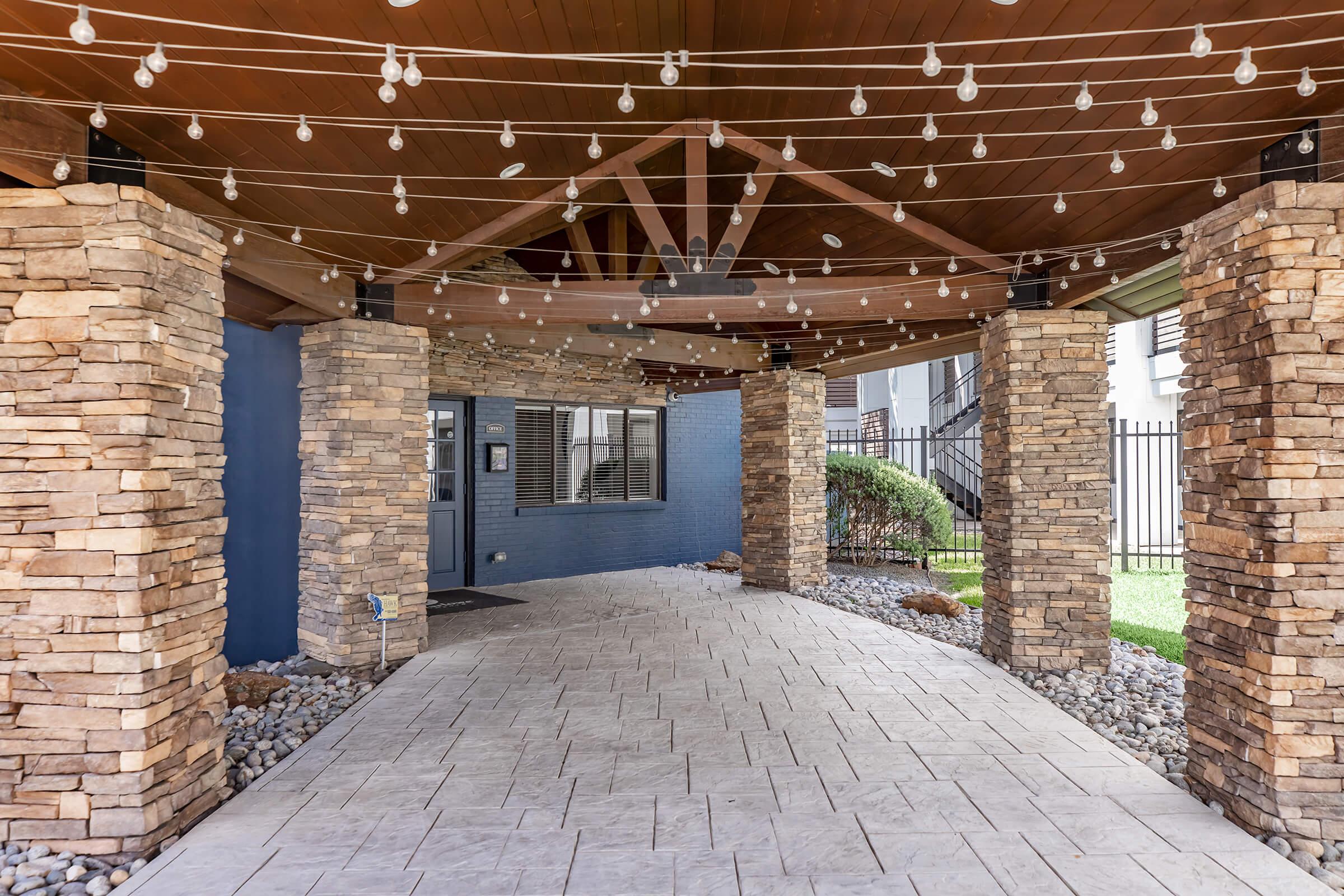 A covered entryway featuring stone columns and decorative string lights. The floor is made of patterned tiles, and there is a blue wall in the background. Lush greenery is visible outside the structure, providing a welcoming atmosphere.