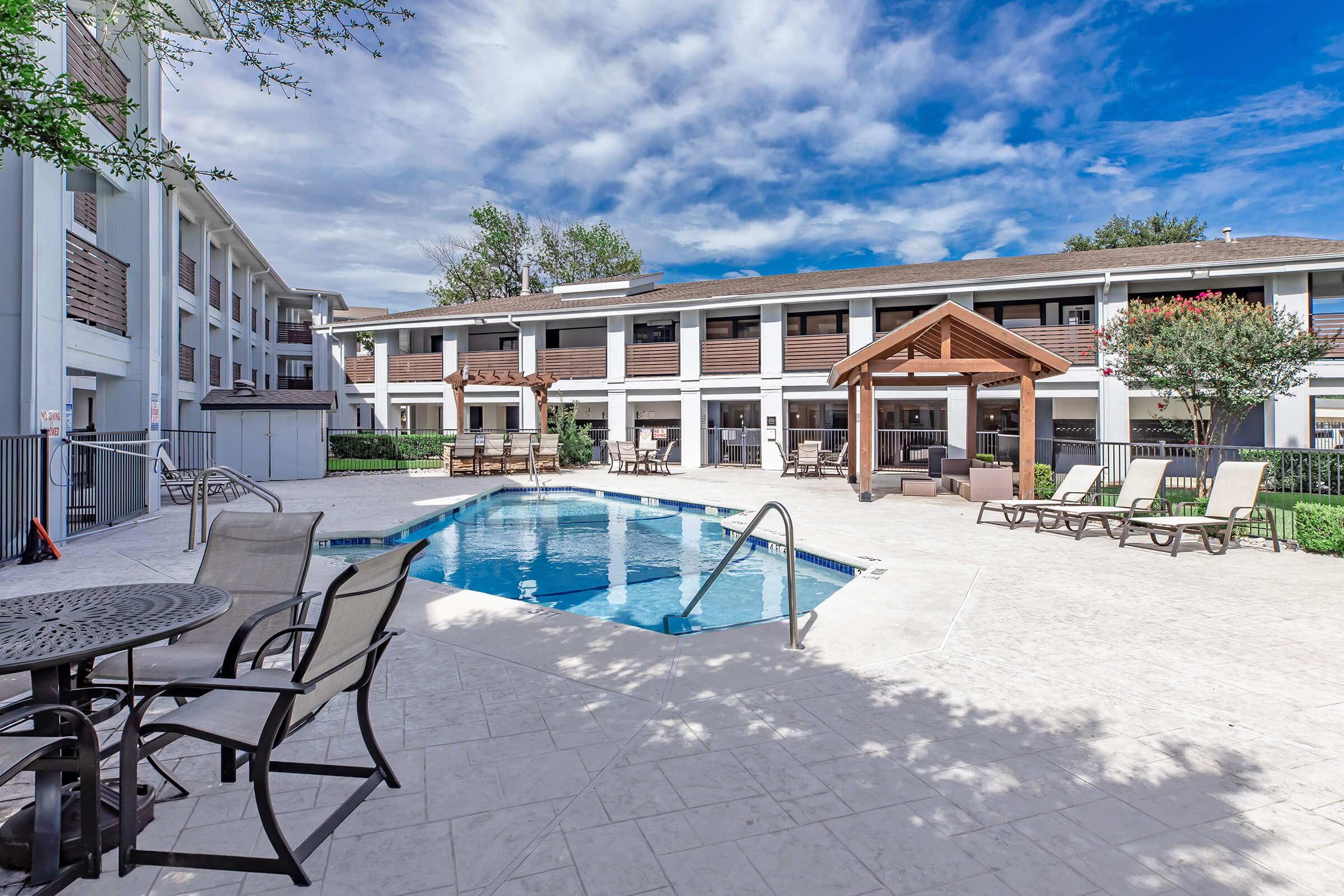 A sunny outdoor pool area at a hotel, featuring lounge chairs and tables. The pool is surrounded by a patio with a decorative gazebo, and several trees provide shade. The building in the background has multiple stories and large windows, creating a welcoming atmosphere. Fluffy clouds decorate the blue sky.