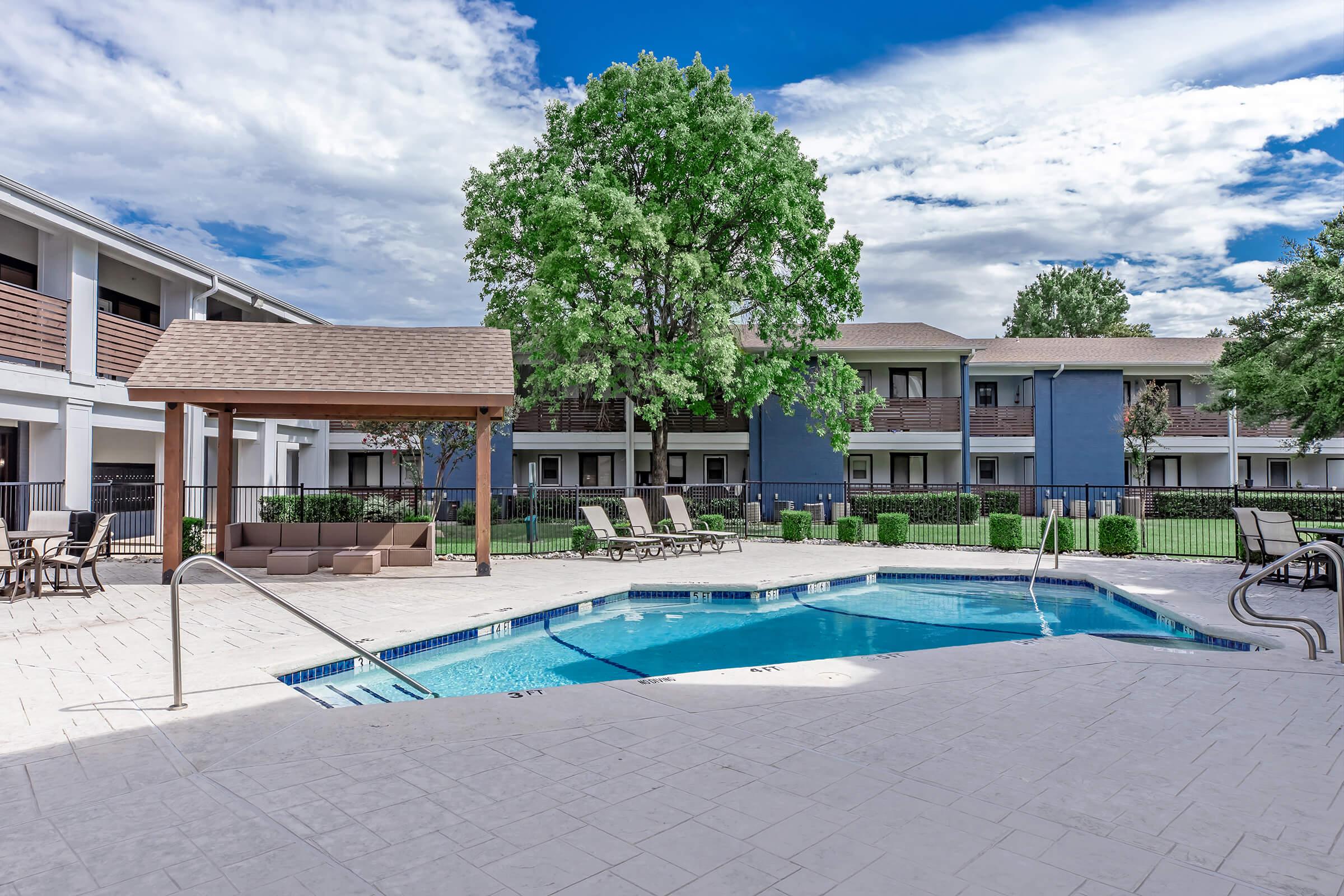 A resort-style outdoor pool surrounded by lounge chairs and a shaded gazebo. Lush landscaping with trees and well-maintained grassy areas in the background. Two-story buildings with balconies are visible, under a partly cloudy blue sky.