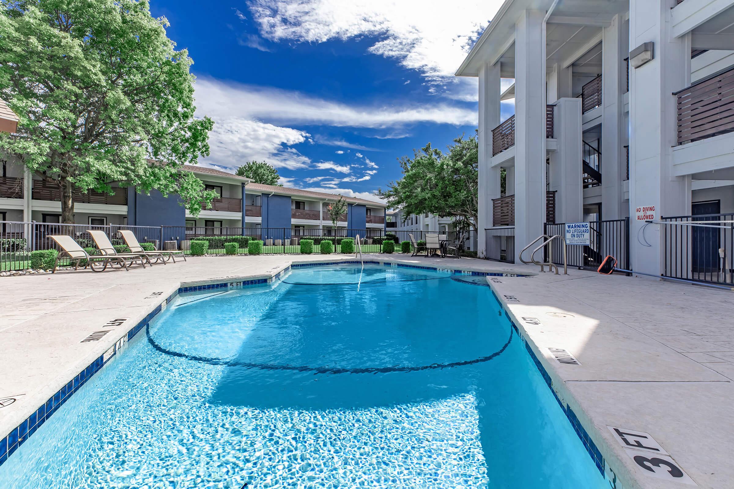 A clear, inviting swimming pool with a fountain feature, surrounded by lounge chairs. The pool area is bordered by a fence and lush greenery, with apartment buildings in the background. The sky is bright blue with fluffy white clouds, creating a pleasant and relaxed atmosphere.