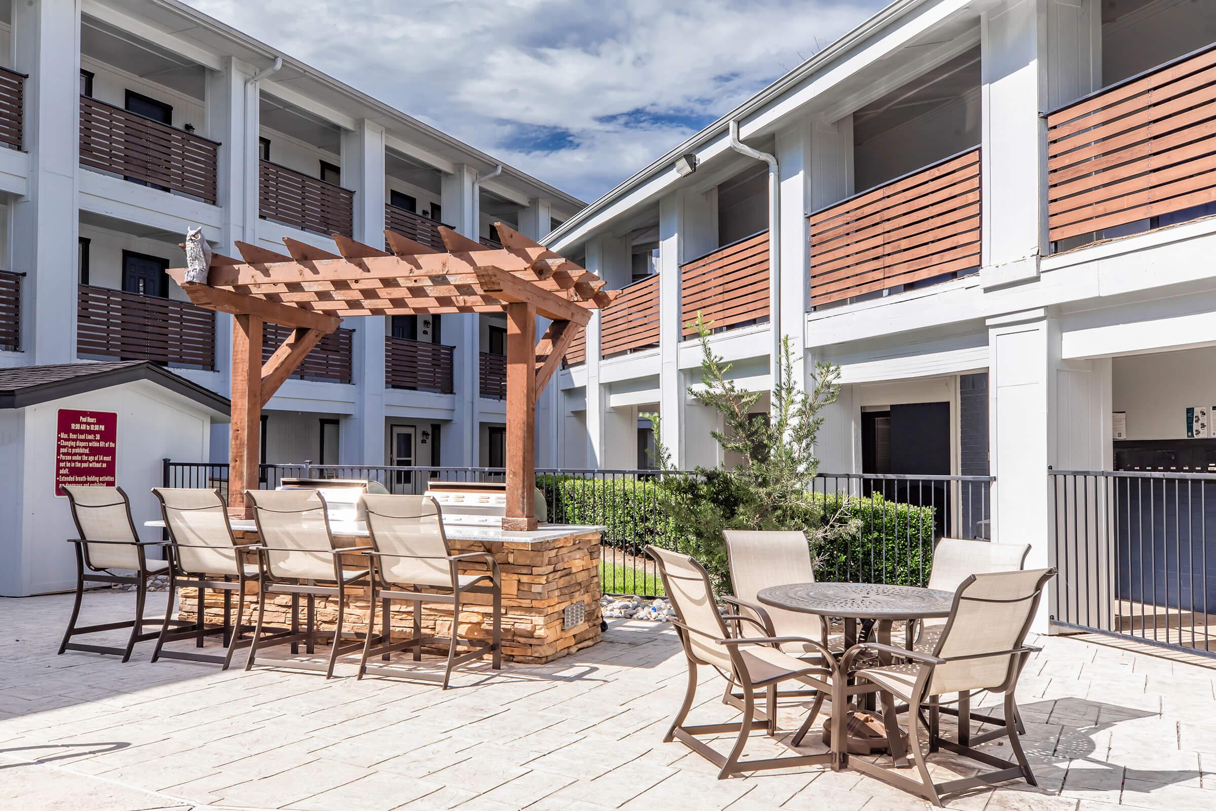 Outdoor seating area with a wooden pergola and stone structure, surrounded by a well-maintained courtyard. The setting includes tables and chairs, with balconies visible in the background. The atmosphere is bright and inviting, ideal for relaxation or social gatherings.