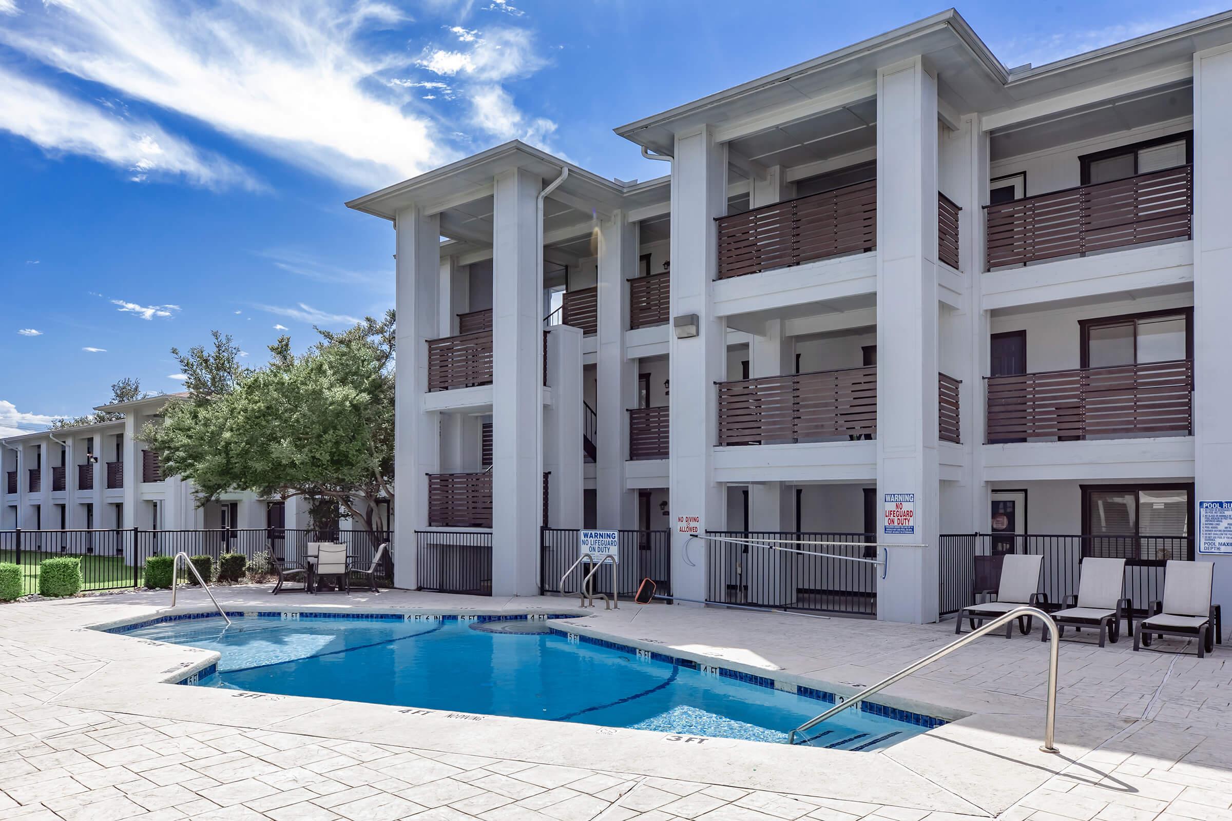 A modern hotel building featuring multiple stories with balconies, overlooking a small swimming pool. The pool area has sun loungers and is surrounded by paved decking. Clear blue skies and a few clouds in the background enhance the relaxing atmosphere.