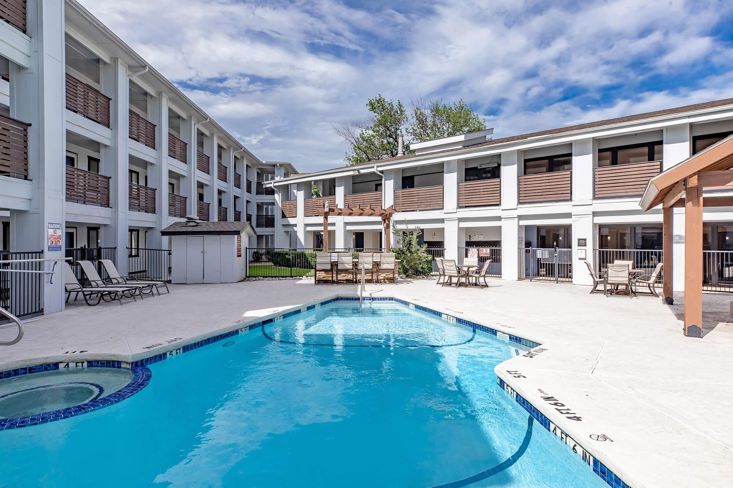 A well-maintained outdoor pool surrounded by lounge chairs and a wooden pergola. The pool area is bordered by two modern buildings with multiple levels. The sky above is partly cloudy, creating a bright and inviting atmosphere for relaxation and leisure.