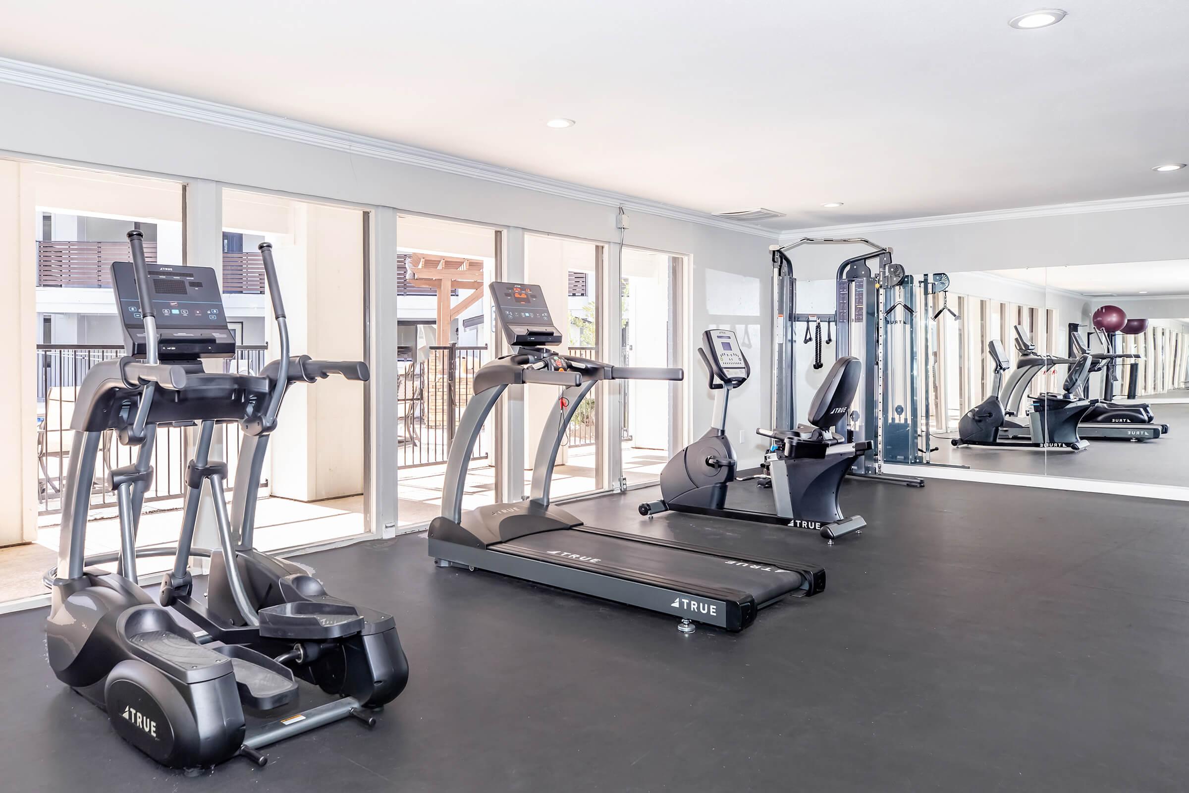 A modern gym interior featuring various exercise equipment including an elliptical machine, a treadmill, and a stationary bike. Large windows allow natural light to fill the space, and mirrors are positioned along one wall. The flooring is dark, providing a sleek, contemporary look.