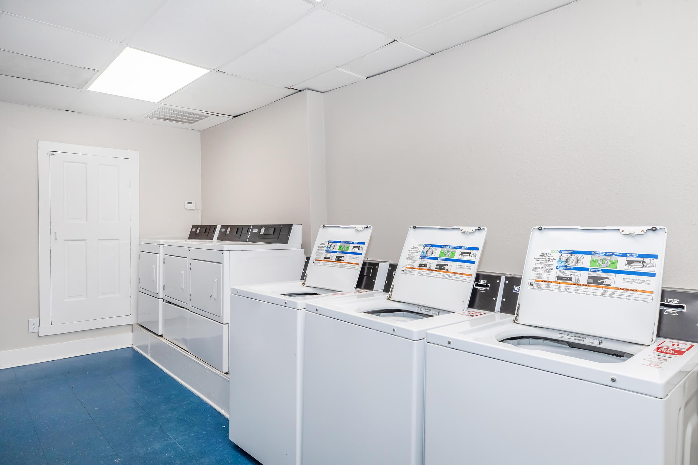A clean and well-lit laundry room featuring three white washing machines with open lids, a row of dryers against the wall, and a simple white door. The floor is blue, and the walls are painted a light color, creating a tidy and functional space for doing laundry.