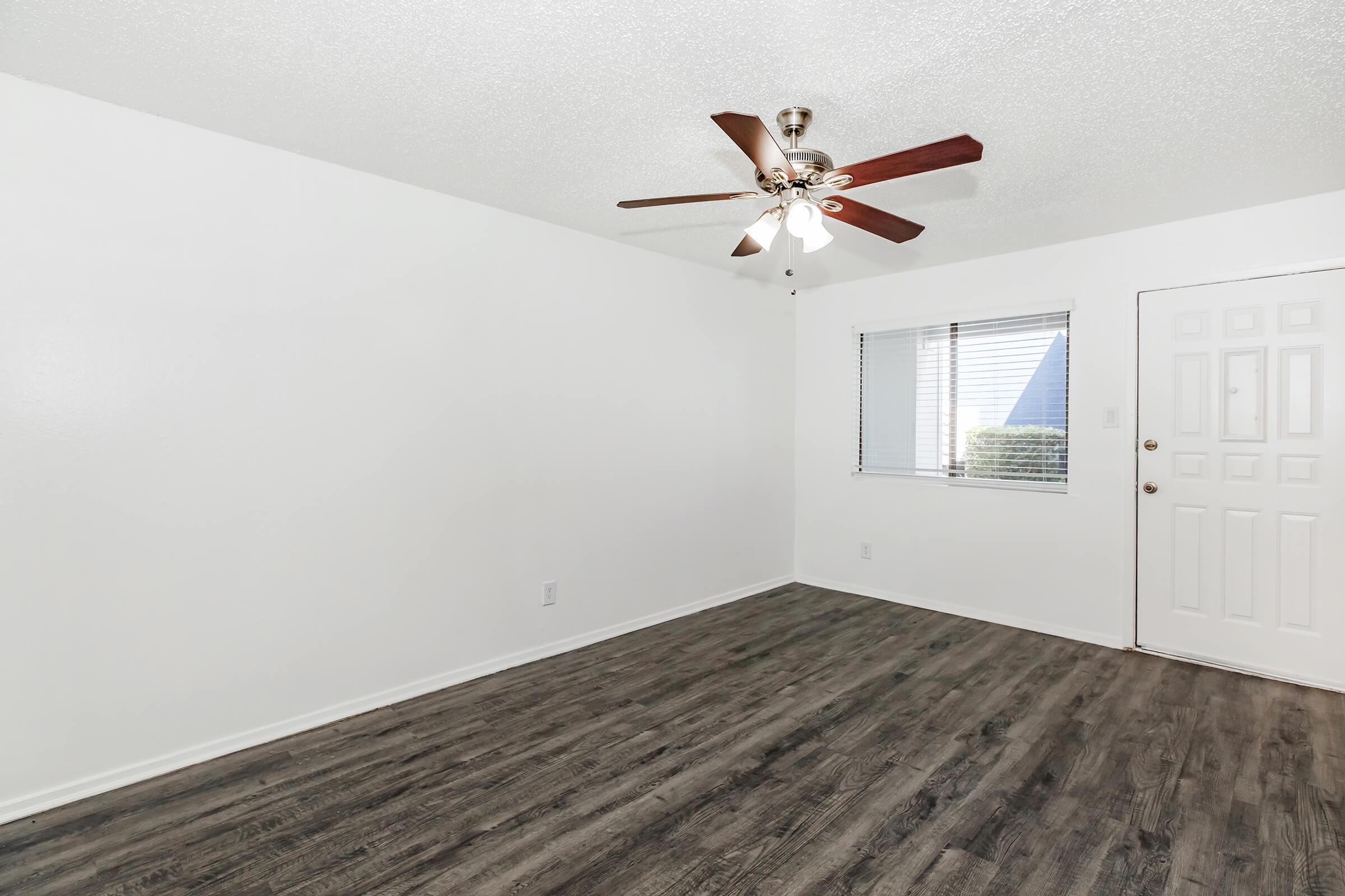 A spacious, empty living room with light-colored walls and a ceiling fan. There is a window with blinds on the left side and a front door on the right. The floor is a dark wood laminate, providing a modern touch to the room. The overall atmosphere is bright and inviting.