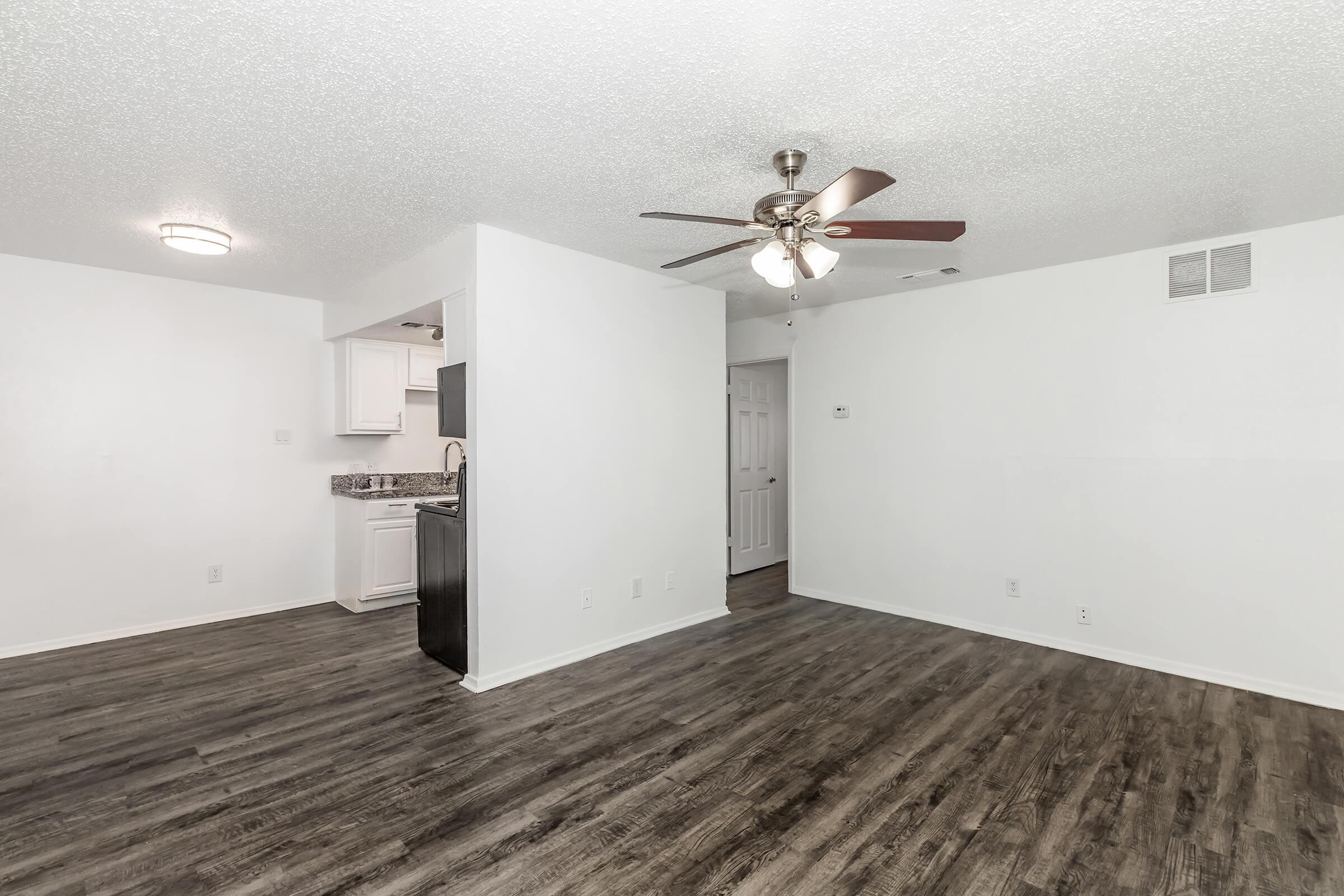 A spacious living area with light-colored walls and a ceiling fan. The room features dark wood-like flooring and an open layout connecting to a kitchen area with white cabinets and a granite countertop. A door leads to another room, and there is ample natural light in the space.