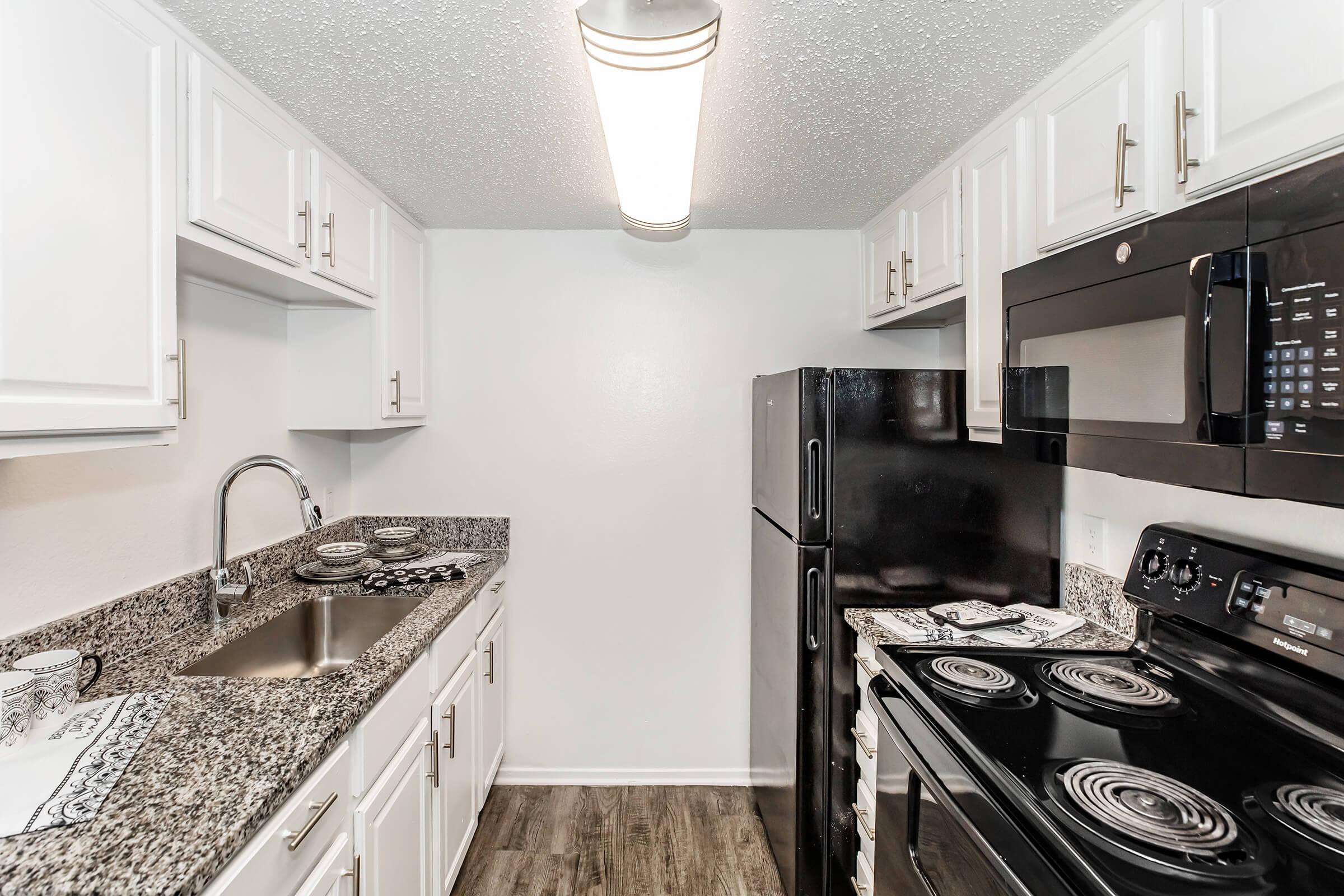 A modern kitchen featuring white cabinets, a granite countertop with a sink, black appliances including a refrigerator, microwave, and stove, and light-colored walls. The space has a clean and organized look, with decorative dishware on the counter and a ceiling light providing illumination.