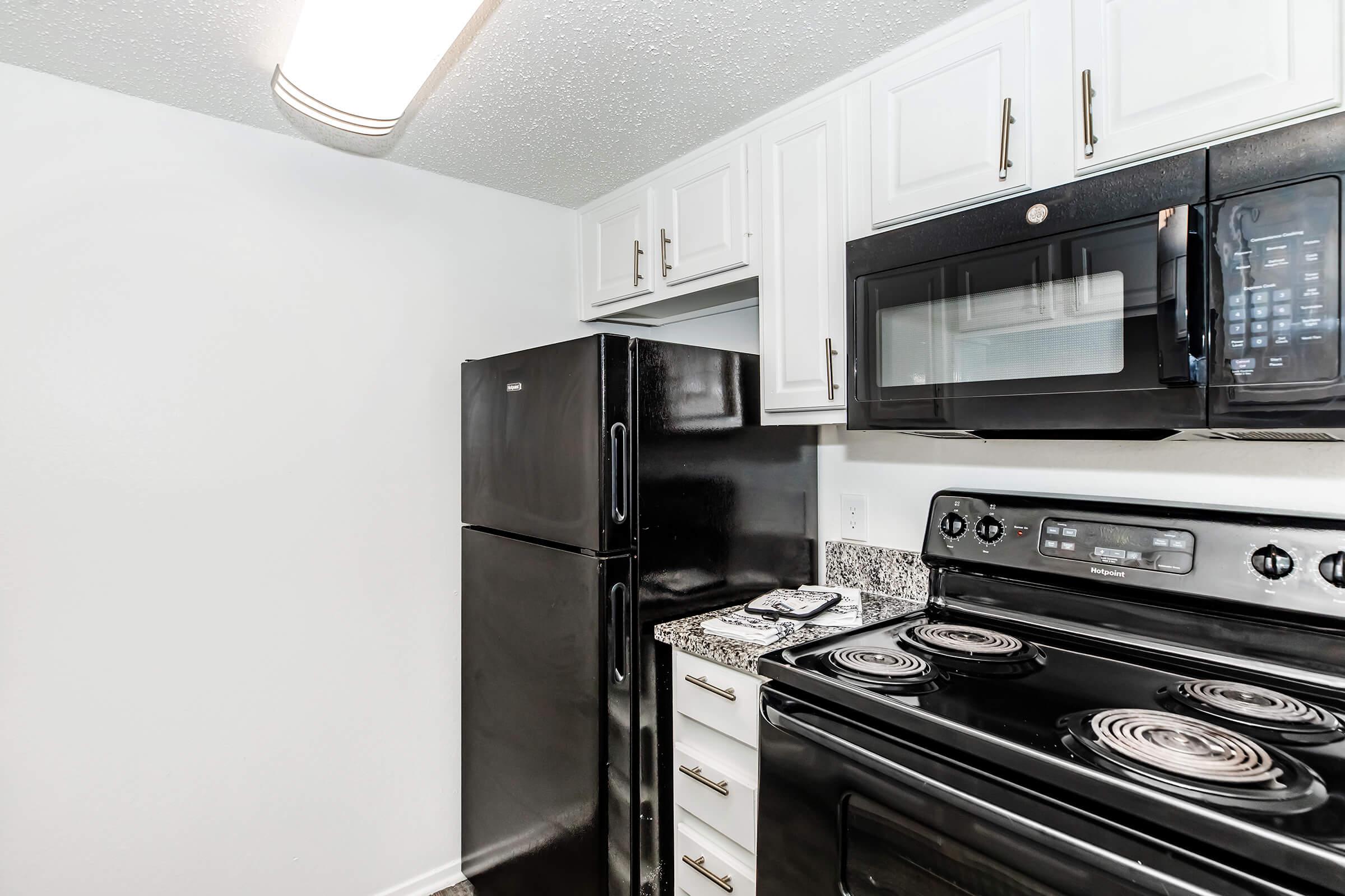 A modern kitchen featuring white overhead cabinets, a black refrigerator, and a black stove with an oven. There is a microwave above the stove, and the countertops are made of granite. The walls are painted white, creating a clean and bright atmosphere.