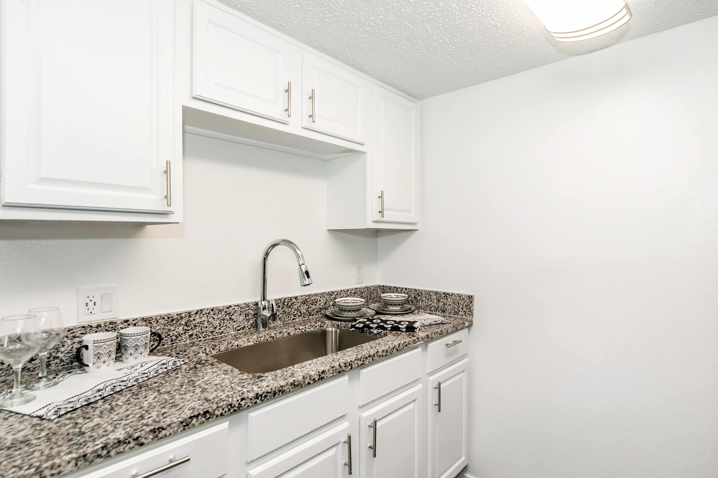 A modern kitchen featuring white cabinetry and a granite countertop. The sink is centrally located with a sleek faucet. There are dishes arranged neatly on the counter, and the space is well-lit with a ceiling light, creating a clean and inviting atmosphere.