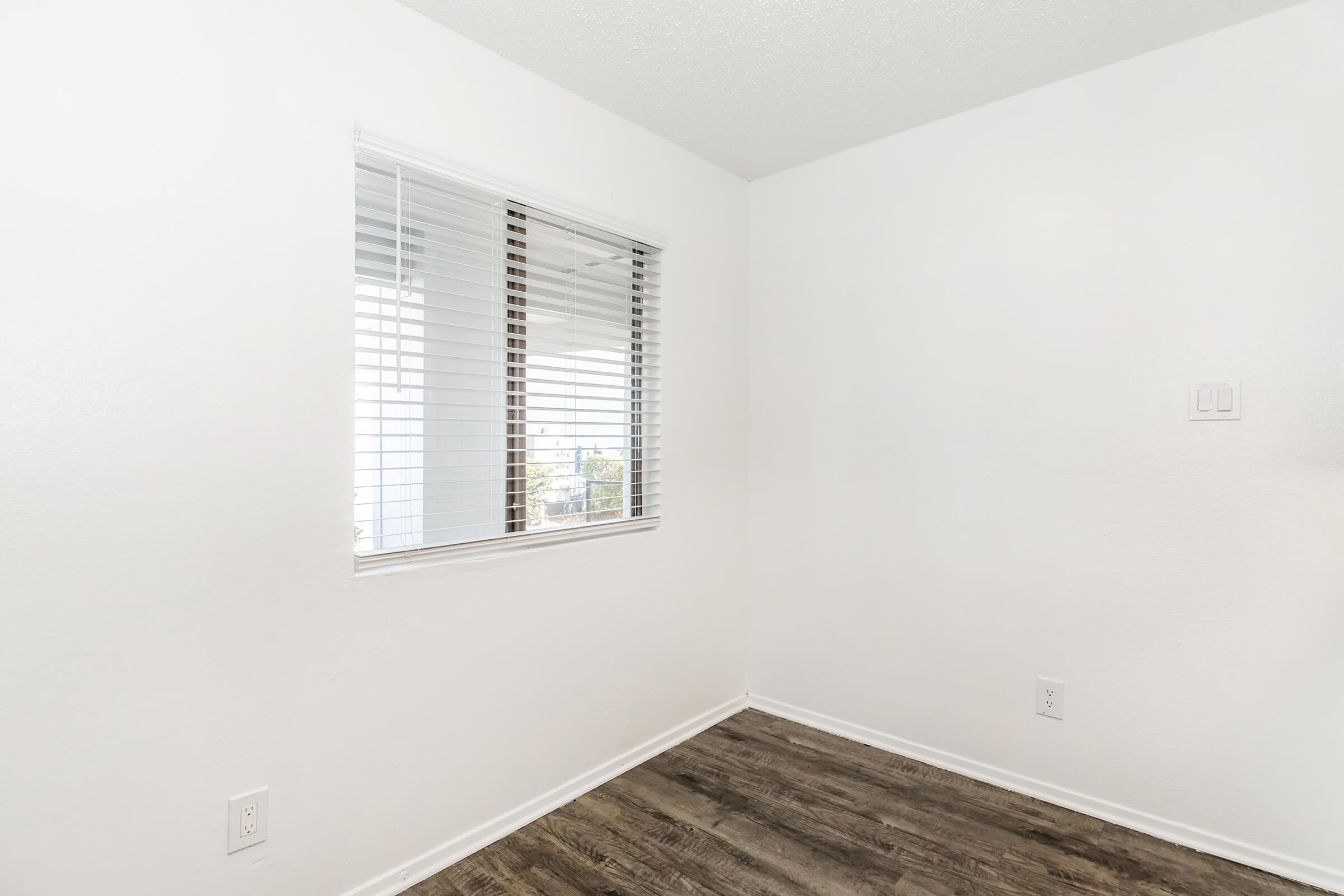 A plain, minimalistic room with white walls and a window featuring white blinds. The floor is covered in a light brown wood-like material. The room appears empty and well-lit, suggesting a clean and modern interior design.