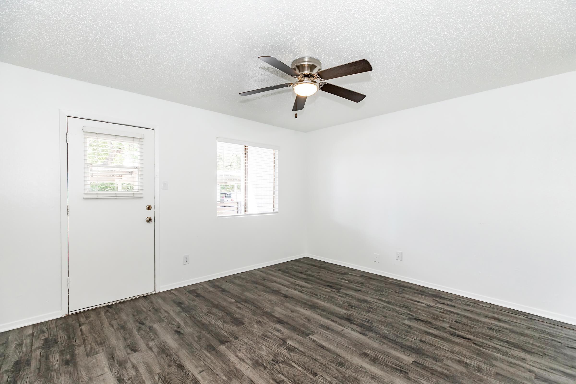 Bright and airy interior space with white walls and a light wood floor. Features include a ceiling fan and two windows allowing natural light, along with a door leading outside. The room appears clean and uncluttered, suitable for various uses.