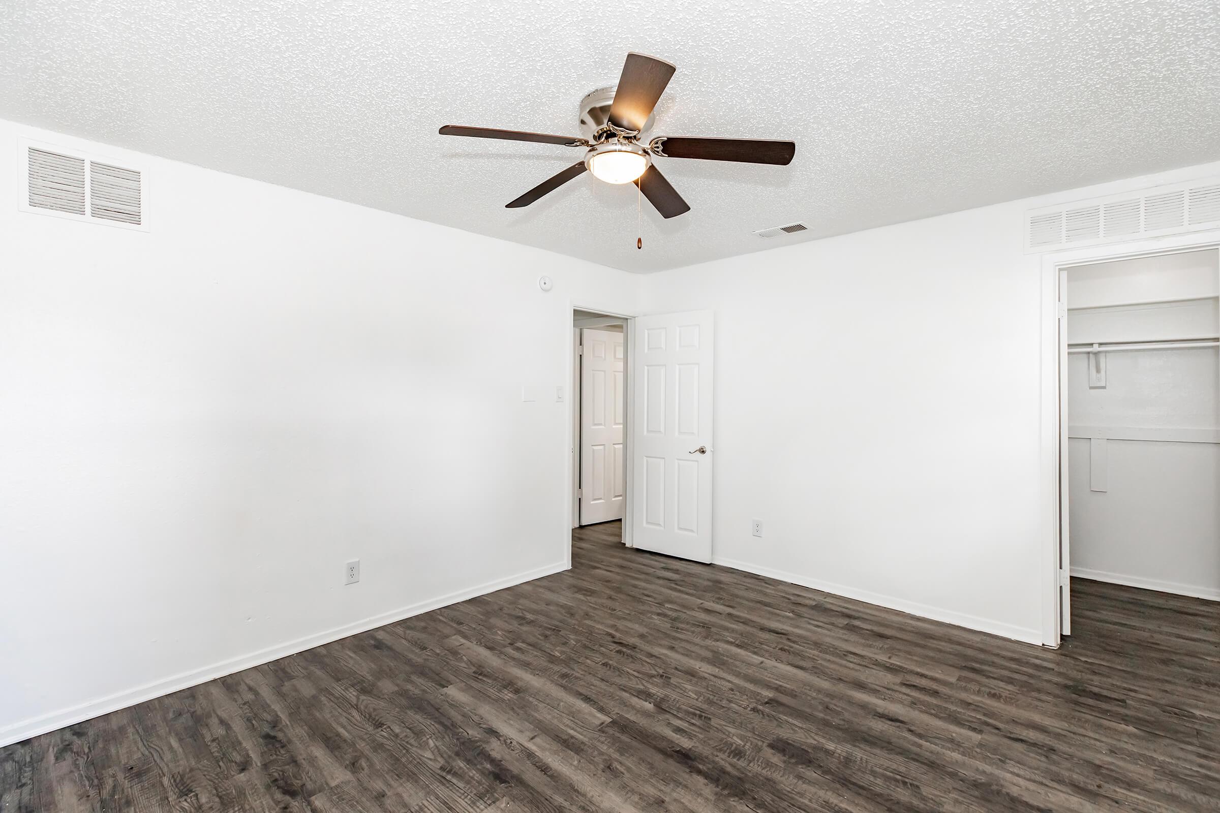 A bright, spacious room with a ceiling fan and light fixture. The walls are painted white, and the flooring is dark laminate. There are two doors leading to additional spaces, one of which appears to be a closet. The overall ambiance is clean and inviting, suitable for various uses.