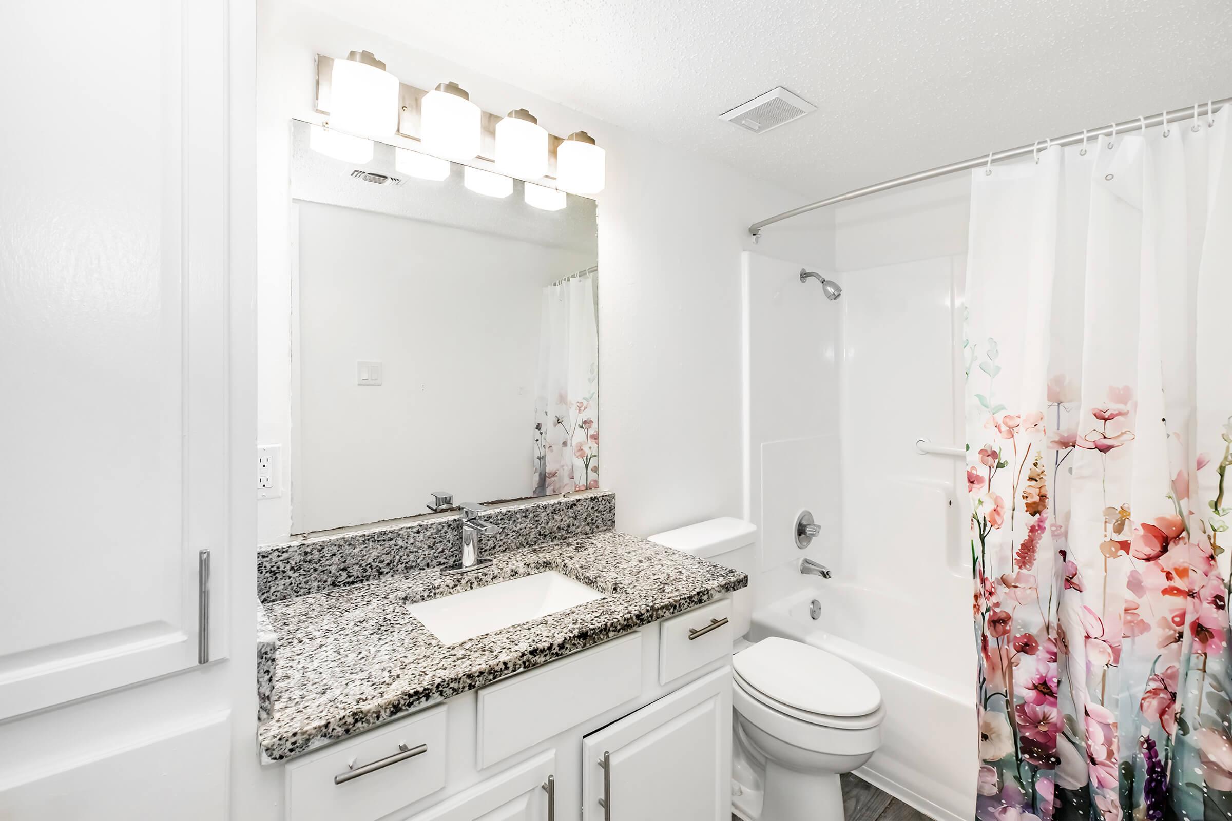 A modern bathroom featuring a white countertop with a sink, a large mirror, and a light fixture above. The shower area has a white bathtub and a floral shower curtain with pink and purple flowers, set against a clean white wall. The floor is completed with neutral-colored tiles.