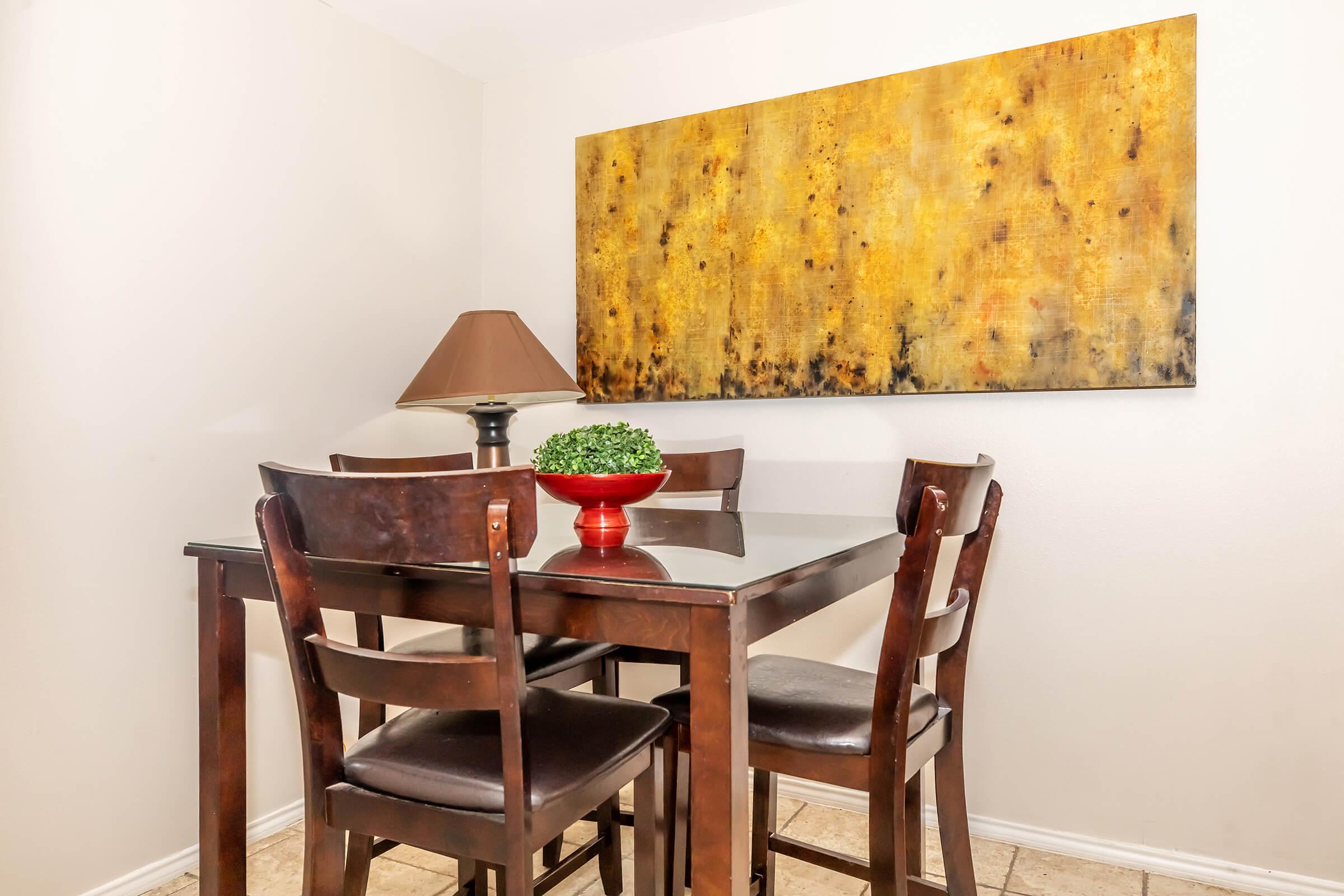 A cozy dining area featuring a dark wooden table with four chairs, a decorative lamp on the table, and a small potted plant. Above the table hangs a large abstract artwork with a golden hue, adding warmth to the space. The walls are light-colored, creating an inviting atmosphere.