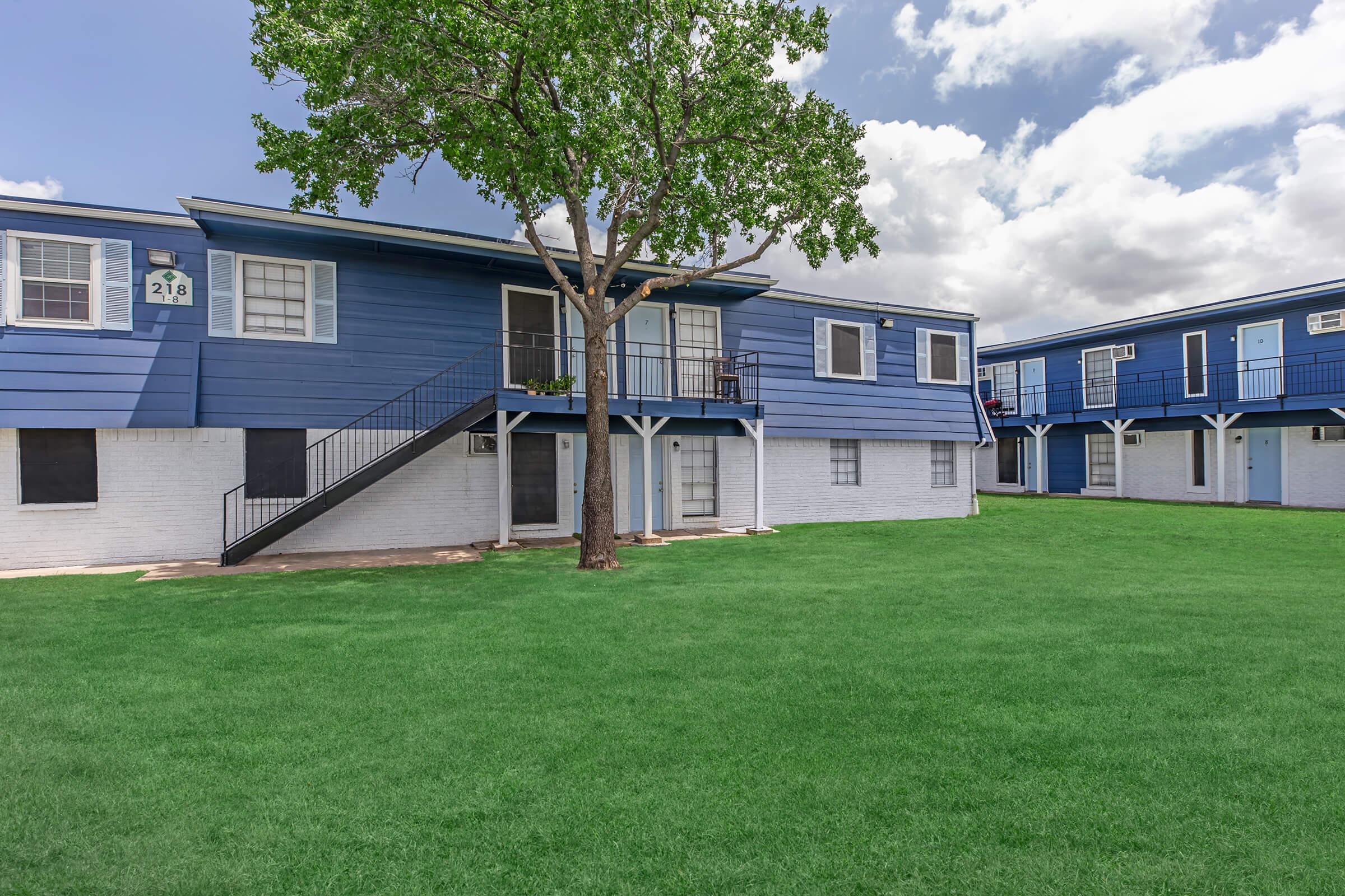 A two-story apartment building with blue siding and white accents, surrounded by green grass. A large tree stands in front of the building. The structure includes staircases leading to the upper level, with balconies visible. The sky is partly cloudy.