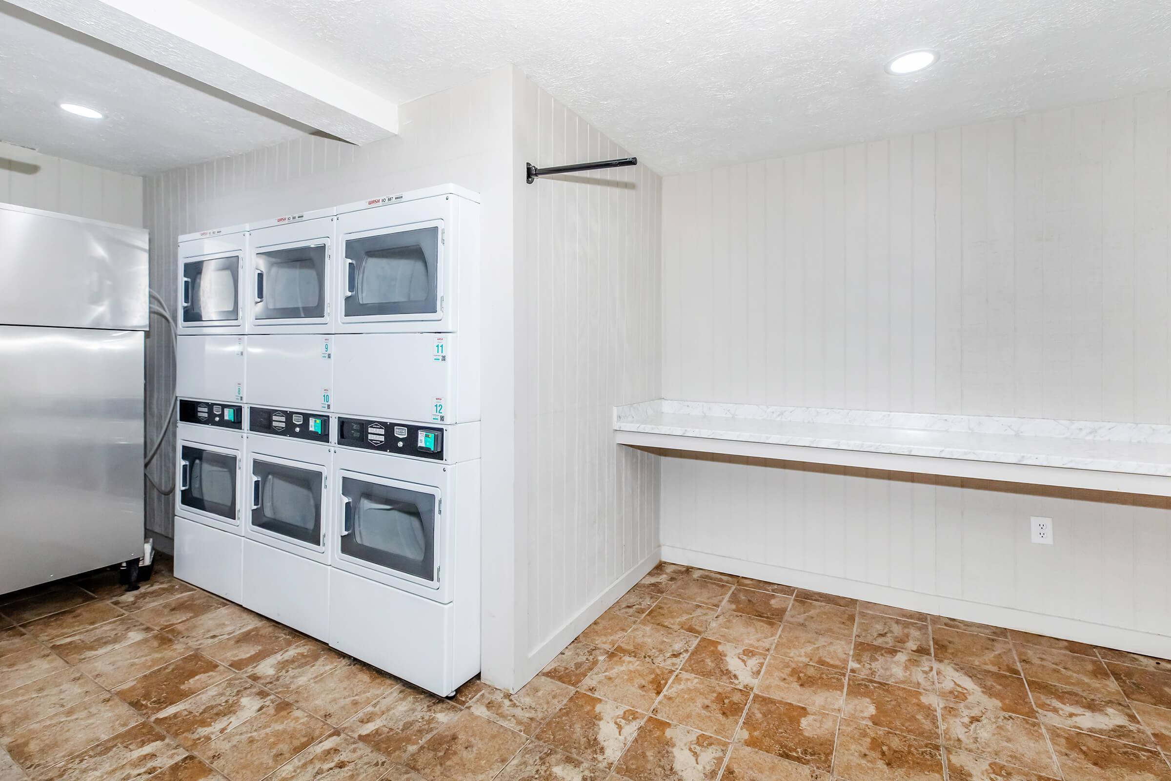 A laundry room featuring stacked washing machines and dryers on the left, a stainless steel refrigerator nearby, and a countertop for folding clothes on the right. The floor is tiled with beige and brown patterns, and the walls are painted in light colors, creating a clean and functional space.