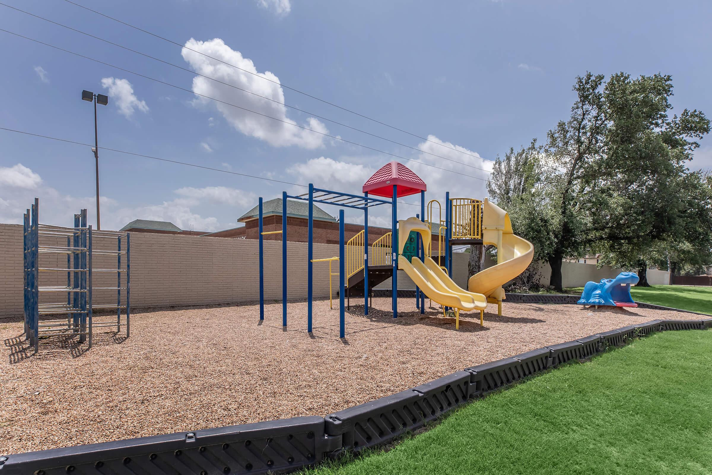 A playground featuring a colorful play structure with a yellow slide, climbing bars, and a blue dinosaur slide, surrounded by wood chips and green grass, under a partly cloudy sky.