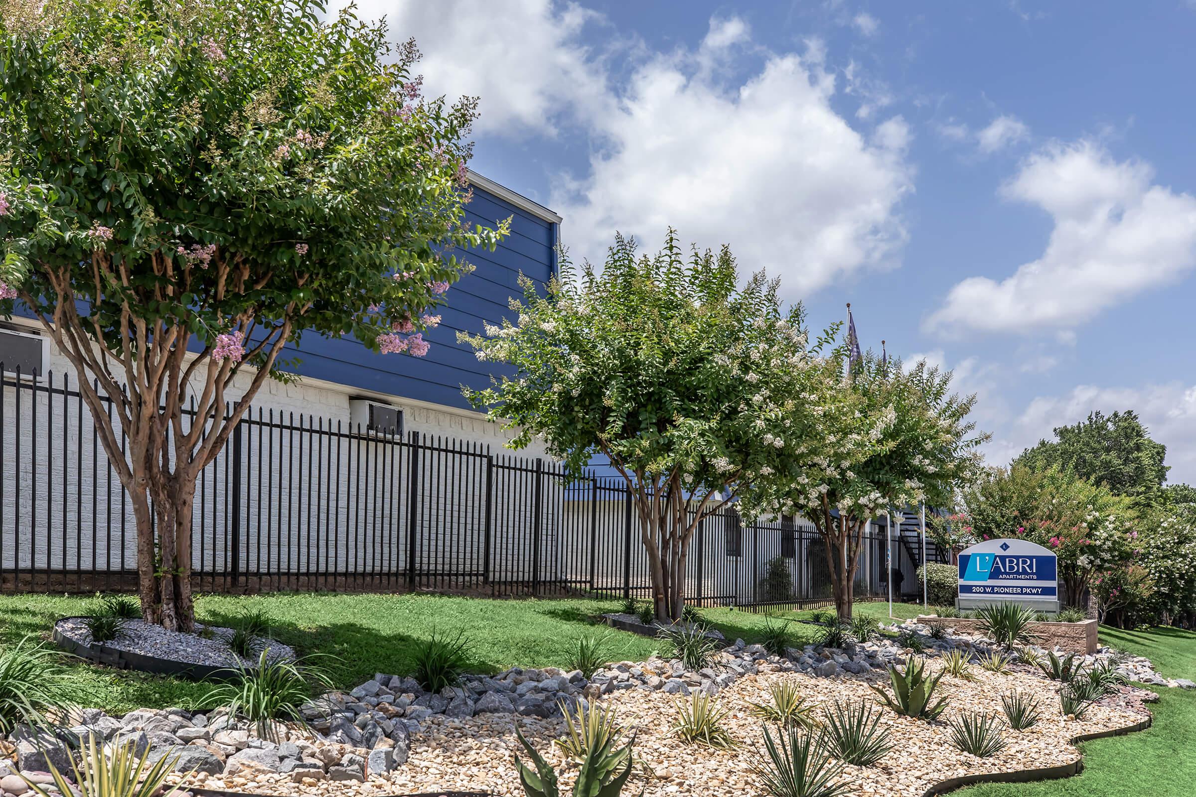 A landscaped area featuring trees and decorative rocks in front of a building with blue walls and a sign that reads "L'Abri." The scene is bright with a partly cloudy sky, showcasing well-maintained greenery.