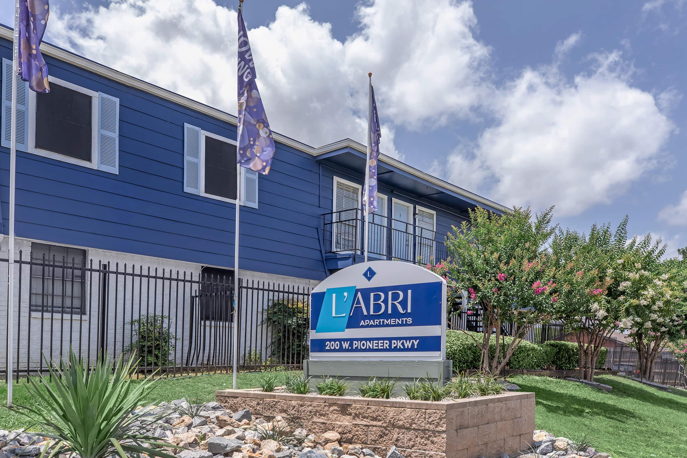 Sign for L'Abri Apartments, featuring the name prominently along with the address "200 W. Pioneer Pkwy." The building in the background is painted blue, and the scene includes flags and landscaped greenery. Fluffy clouds are visible in the sky above.