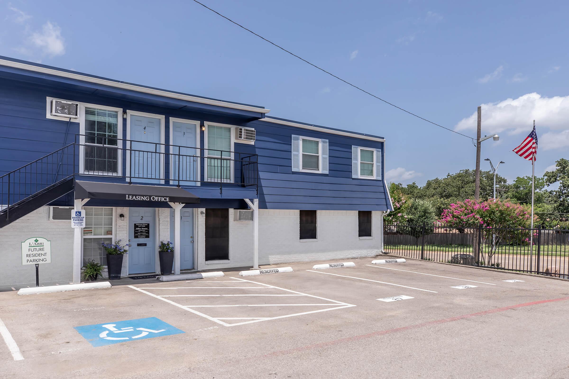 A blue and white leasing office building with a sign that reads "Leasing Office." There is a parking area with a disabled parking space visible, and in the background, a flagpole with an American flag. The scene is bright and sunny, with trees in the vicinity.