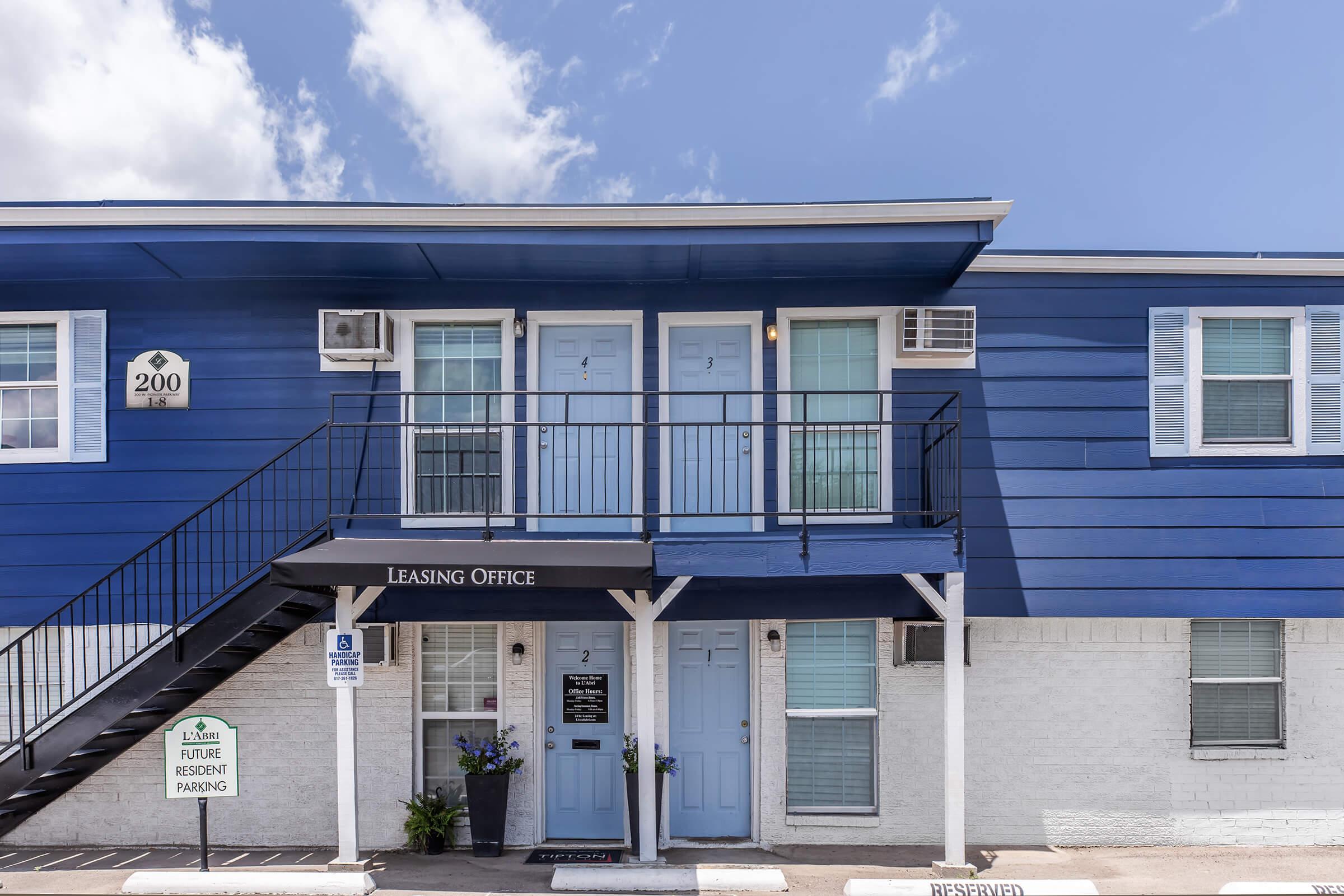 A blue two-story building with a leasing office. The office has a black awning labeled "Leasing Office" and features two light blue double doors. Stairs lead to the second floor, and there are signs for reserved parking and future resident parking. The sky is clear with a few clouds.