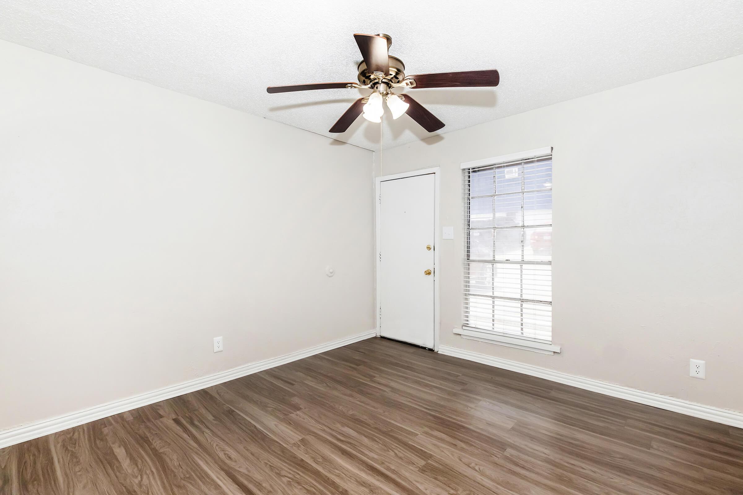 A bright, empty room with light-colored walls and a ceiling fan. There is a closed door with a window beside it, allowing natural light to enter. The floor features laminate wood, contributing to a modern and clean look.