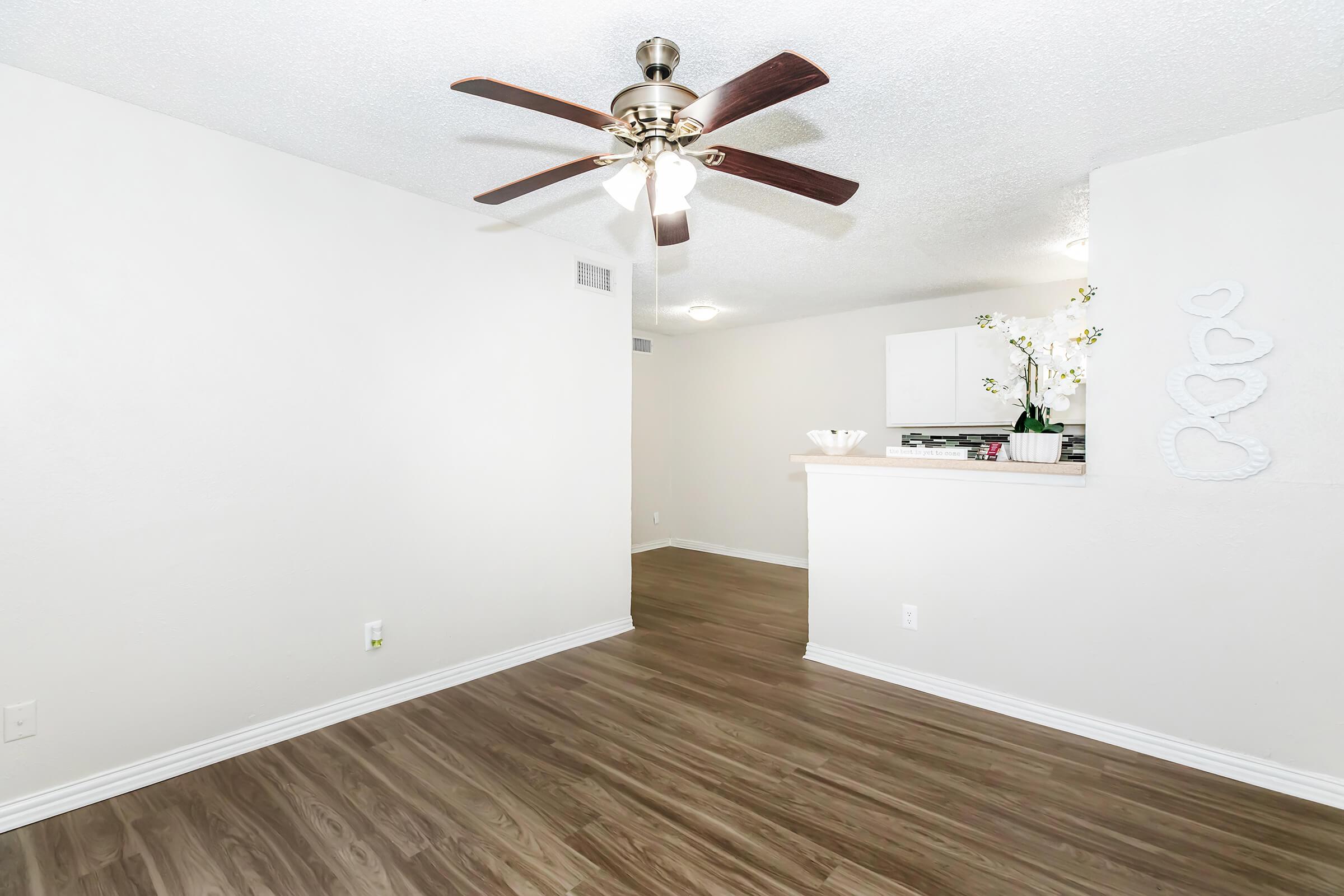 A well-lit room featuring light-colored walls and a wooden floor. A ceiling fan with wooden blades is visible in the center. There is a small bar area separating the living space from a kitchen, which has white cabinetry. Decorative elements include a vase of flowers and a heart-shaped wall decoration.