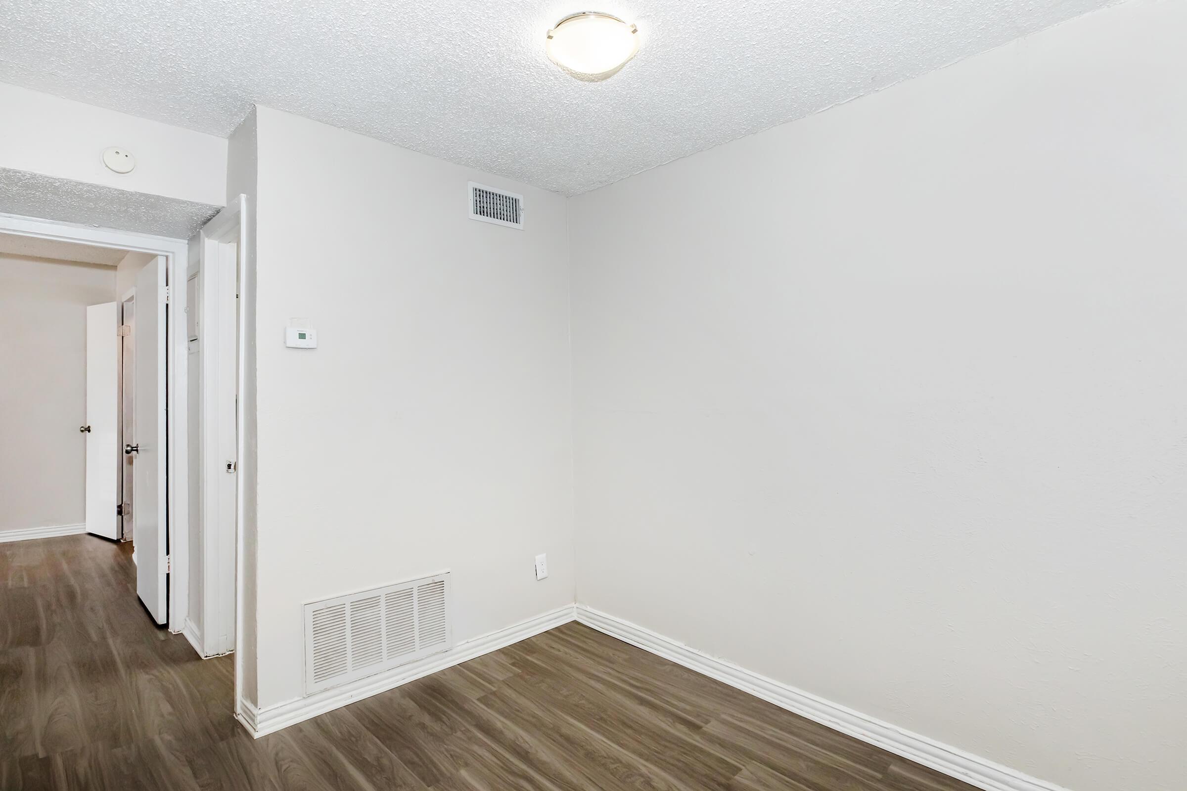 Empty room featuring a light fixture on the ceiling, beige walls, and wooden flooring. There are doorways leading to adjacent rooms, and a wall vent is visible. The overall space is bright and uncluttered, providing a blank canvas for decoration or furnishing.